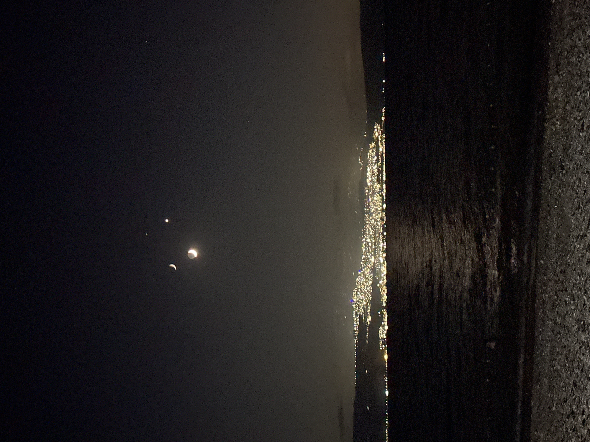 The image depicts a nocturnal landscape over the Sea of Galilee, with the lights of a city in the Emek HaYarden Regional Council, Israel, illuminating the horizon. Three celestial bodies are visible in the dark sky: a crescent moon, a brighter, fuller moon, and a small, distinct point of light, likely a planet or bright star. The sea is dark with subtle reflections of the city lights. The foreground shows a pebbled shoreline. There are no people or animals visible in the frame.