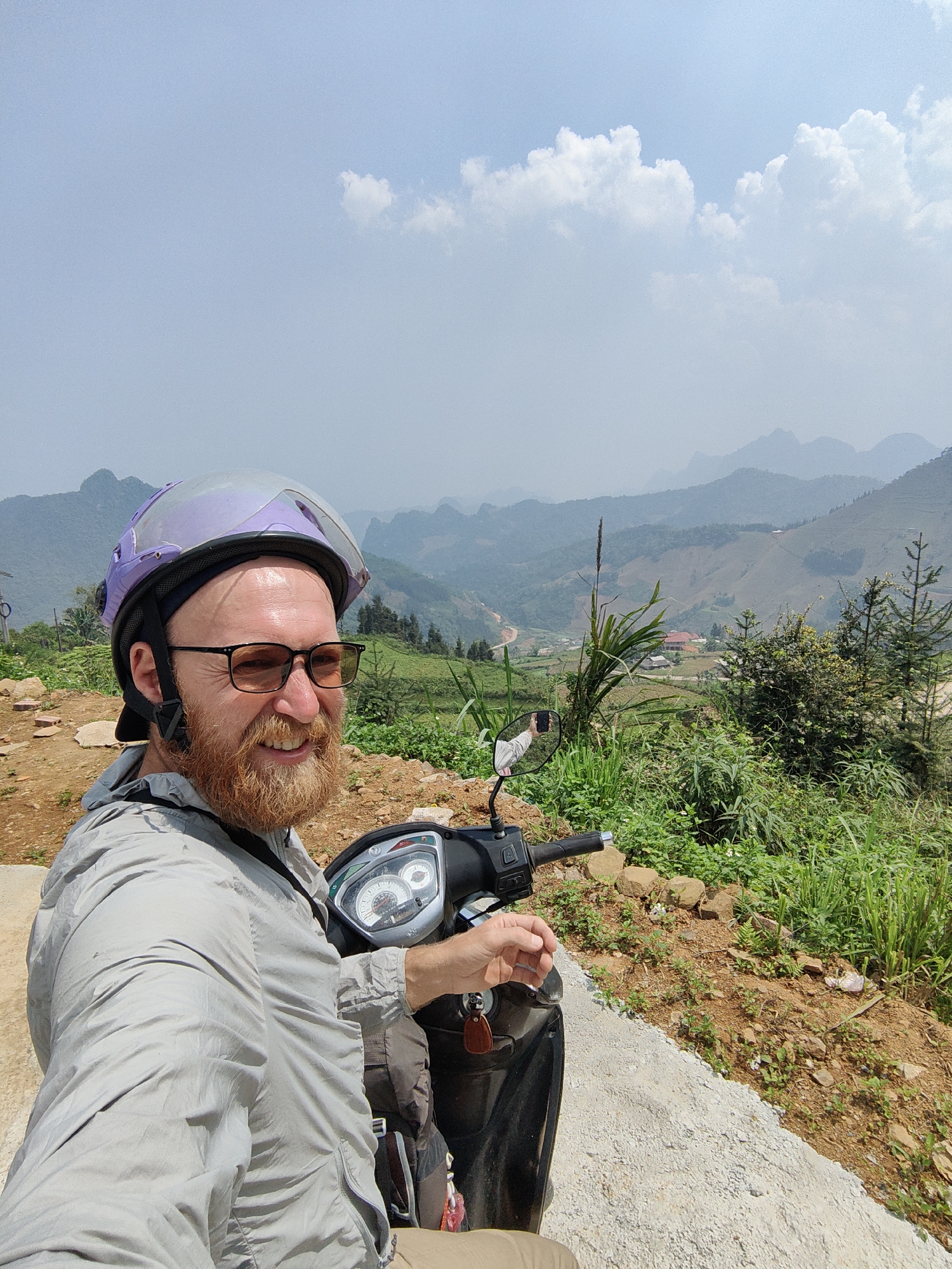 A man wearing a helmet and glasses is sitting on a scooter in Xã Cần Yên, Vietnam. He has a beard and is wearing a light-colored jacket. The scooter is parked on a concrete edge. Behind him is a landscape of green hills and mountains under a partly cloudy sky. Some trees and vegetation are visible nearby.