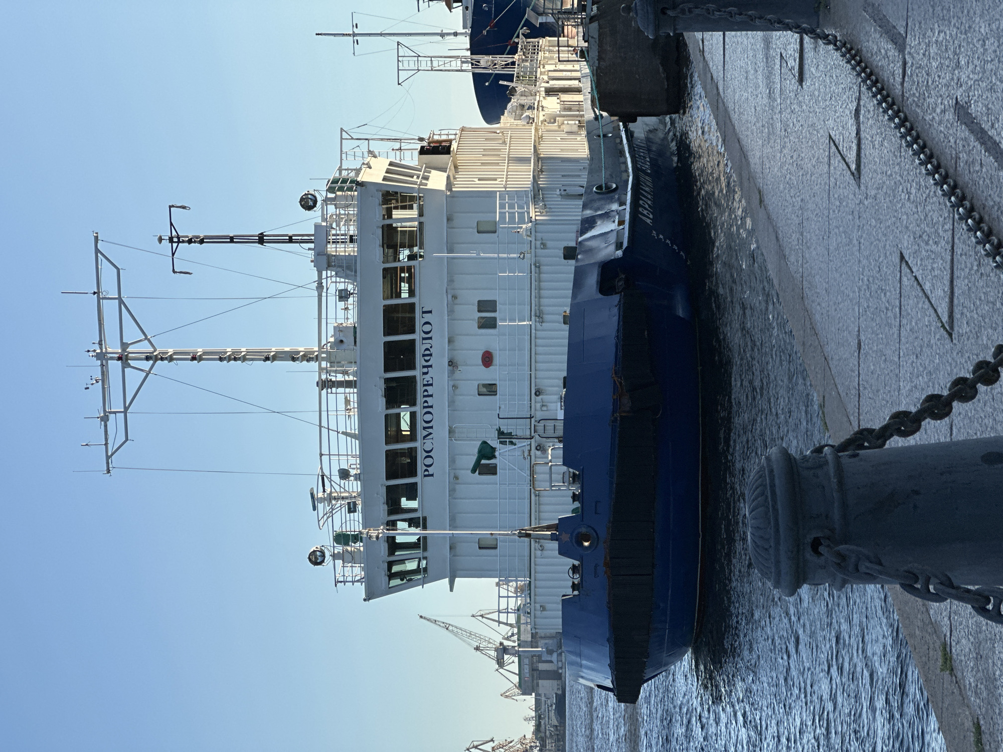 The image shows a large white and blue vessel docked in a city harbor under a clear blue sky. The vessel has "РОСМОРРЕЧФЛОТ" written in blue letters on its upper structure. In the background, several cranes are visible, suggesting this is a port or shipyard. The foreground features a stone walkway with chains and bollards, leading to the edge of the water where the ship is moored. The water appears calm. The overall scene depicts a working port environment with a prominent vessel as the central subject. This location is likely the Saint Petersburg Port, also known as the Big Port of Saint Petersburg, located in Saint Petersburg, Russia.