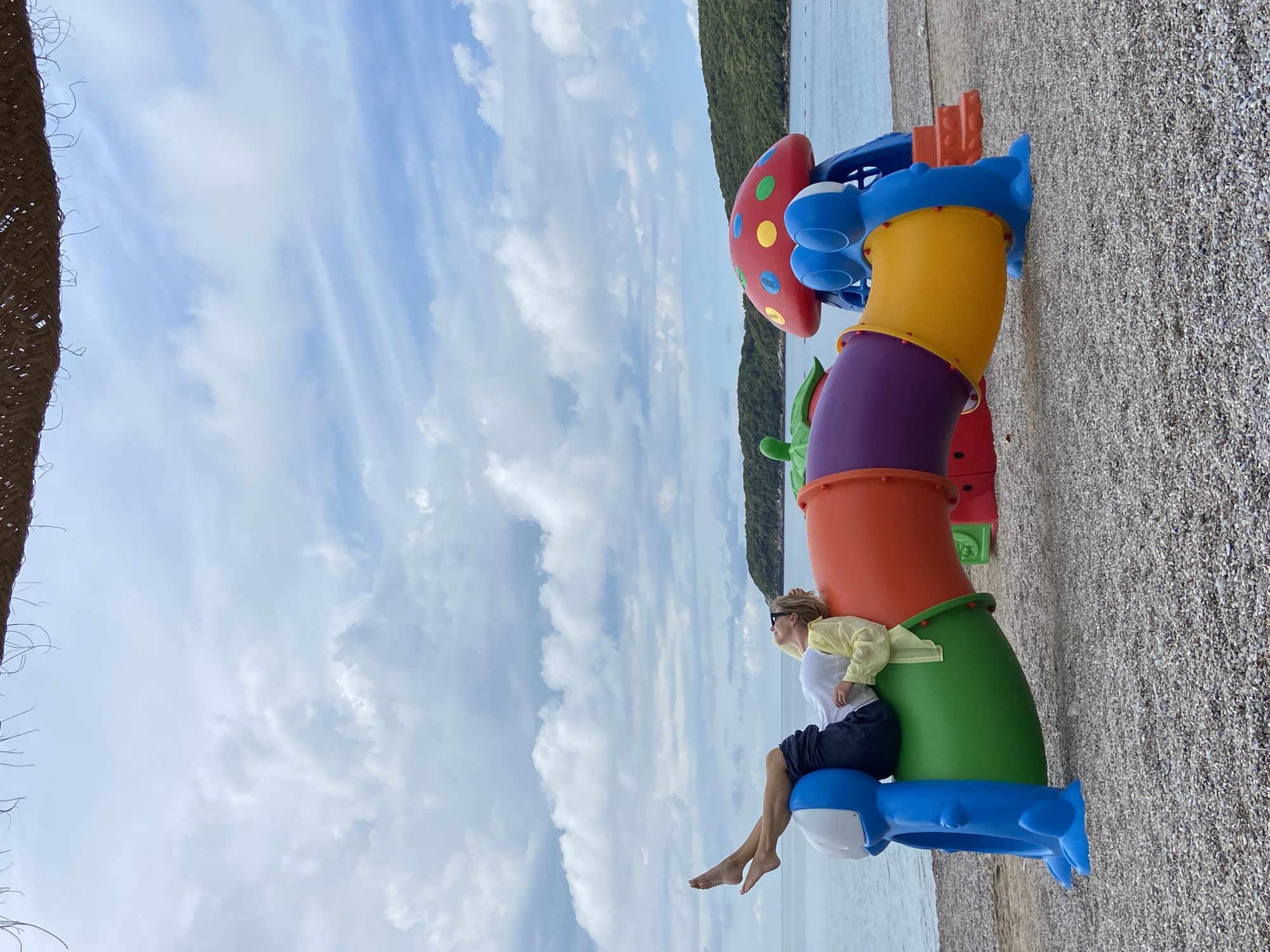 A person is sitting on a colorful children's play structure on a pebbled beach in Prijevor, Montenegro. The play structure is composed of a series of interconnected, tubular sections in green, orange, purple, and yellow, topped with a mushroom-shaped red structure with colorful spots. The person is sitting on a blue section shaped like a whale, near the bottom of the structure. They are wearing a white shirt, dark shorts, and a light yellow jacket, and appear to be gazing out at the water. The sky is partly cloudy and there is a wooded shoreline in the background. A portion of a thatched roof is visible in the upper left corner of the frame.