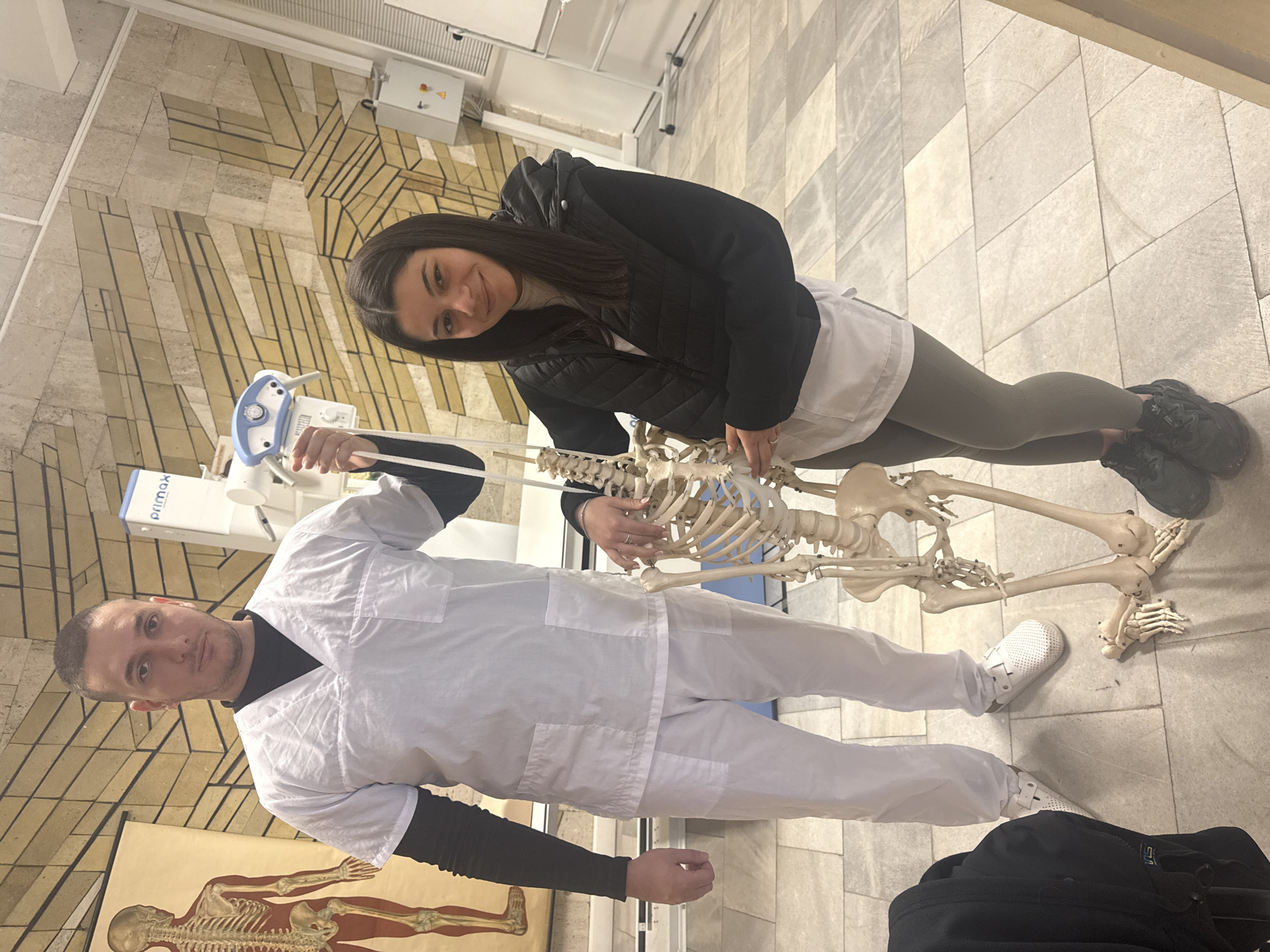 Two people are in a room with a human skeleton. A woman, wearing a black jacket, white shirt, and gray leggings, stands to the right of the skeleton and poses while touching the rib cage. A man, wearing a white lab coat, stands to the left of the skeleton and looks towards the camera. The skeleton stands between them.
In the background, there is what appears to be medical or anatomical equipment. The floor is tiled. A poster with an anatomical illustration is visible near the man. The background has stone and brick architectural elements, possibly in Pleven, Bulgaria, given the prompt.