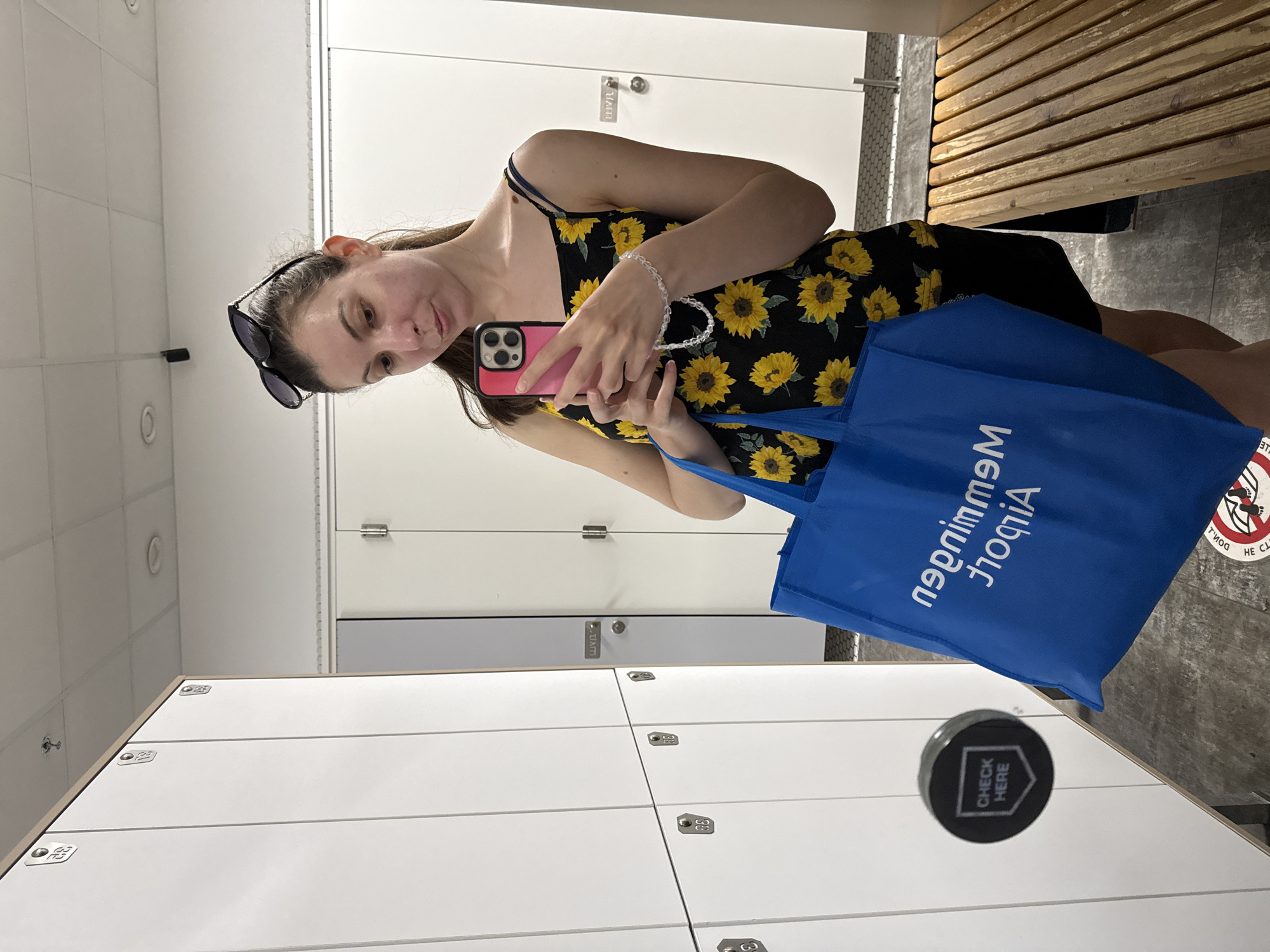 A young woman is standing in a changing room with white lockers in the city of Unknown, Unknown. She is holding a pink cell phone in her right hand and a blue bag in her left. She is wearing a sunflower patterned dress and sunglasses on her head. A wooden bench is visible in the background. A "Check Here" sticker is on the locker in the foreground. The text "Mallorca Memmingen" is printed on the blue bag.