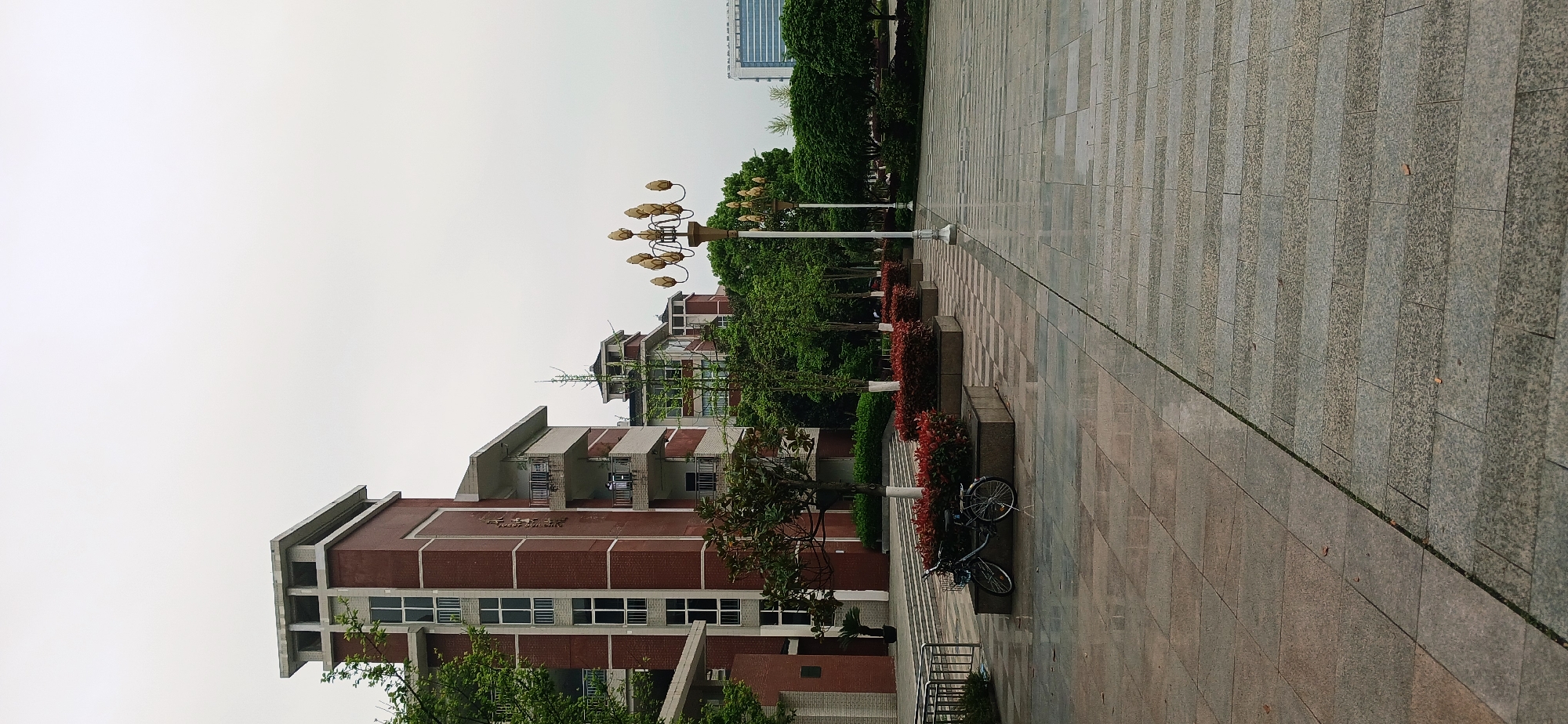 The image shows a street scene in Chongqing High-tech Zone, China. The foreground features a paved walkway of grey stone leading into the distance. Adjacent to the walkway is a raised flowerbed with red foliage. A row of streetlights with decorative lotus-shaped fixtures stands alongside the flowerbed. Further back, red brick buildings with white-framed windows and projecting rooflines are visible. Two parked bicycles are situated next to the flowerbed and buildings. The sky overhead is overcast and grey. In the distant background, a tall skyscraper can be faintly seen. The photo appears to be taken at ground level.