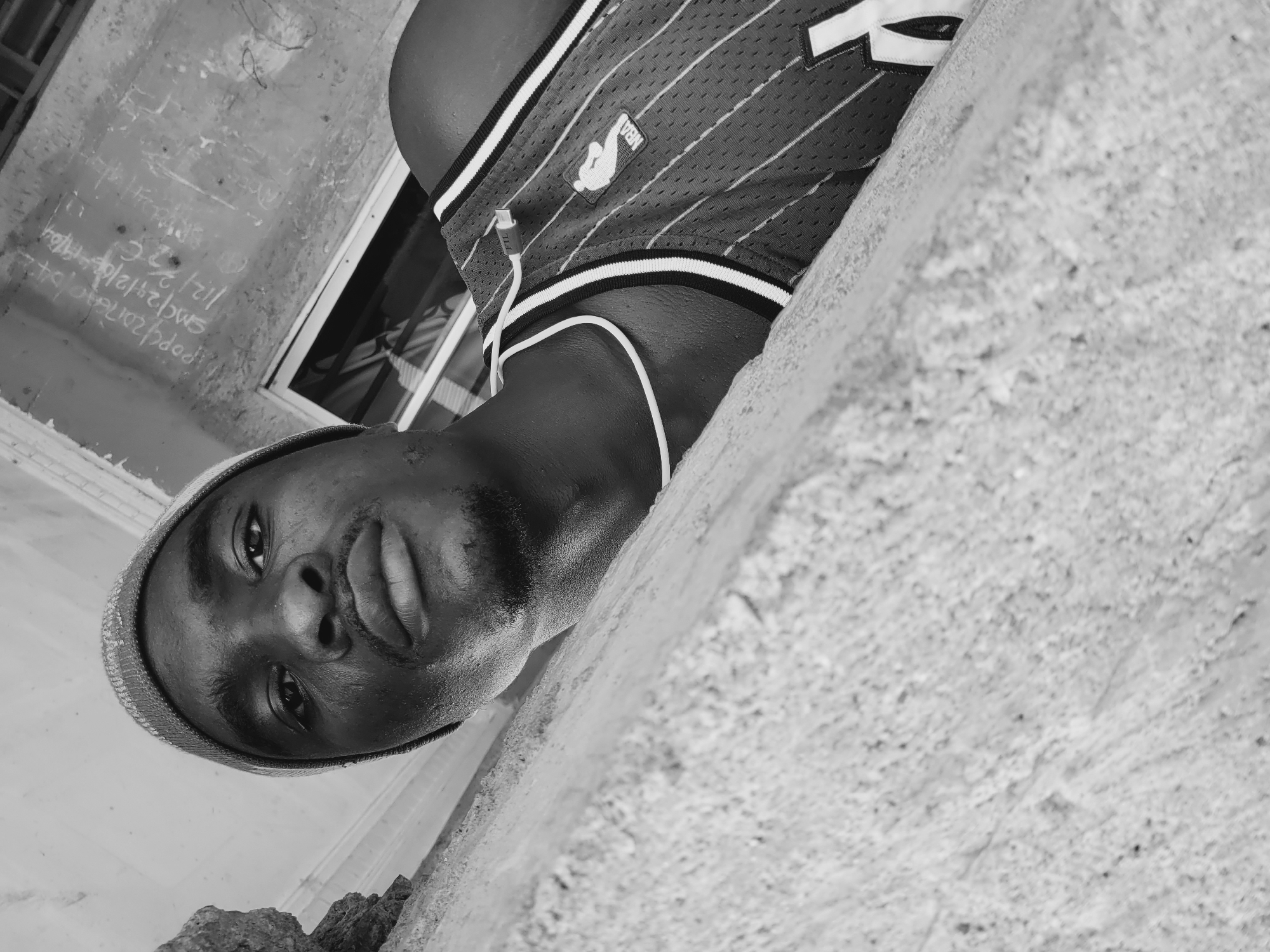 The image is a black and white, low-angle selfie of a young Black man wearing a beanie and an NBA jersey. He is leaning against a textured, light-colored wall, possibly concrete, which occupies the lower portion of the frame. The man's face is partially obscured by the wall, and he is looking directly at the camera with a neutral expression. His left ear is visible, and he appears to have a mustache and a goatee. He is wearing a beanie with a textured pattern and an NBA jersey with white pinstripes and trim. A white cord, likely from headphones, is visible around his neck and plugged into a small device on his jersey. In the background, a window with a black frame and horizontal bars is visible, and a wall with chalk or paint markings is present. The markings appear to include dates and text that are partially obscured and difficult to decipher. The overall setting suggests an outdoor urban environment. Given the provided location, it is implied this scene is in Bukuru, Nigeria.