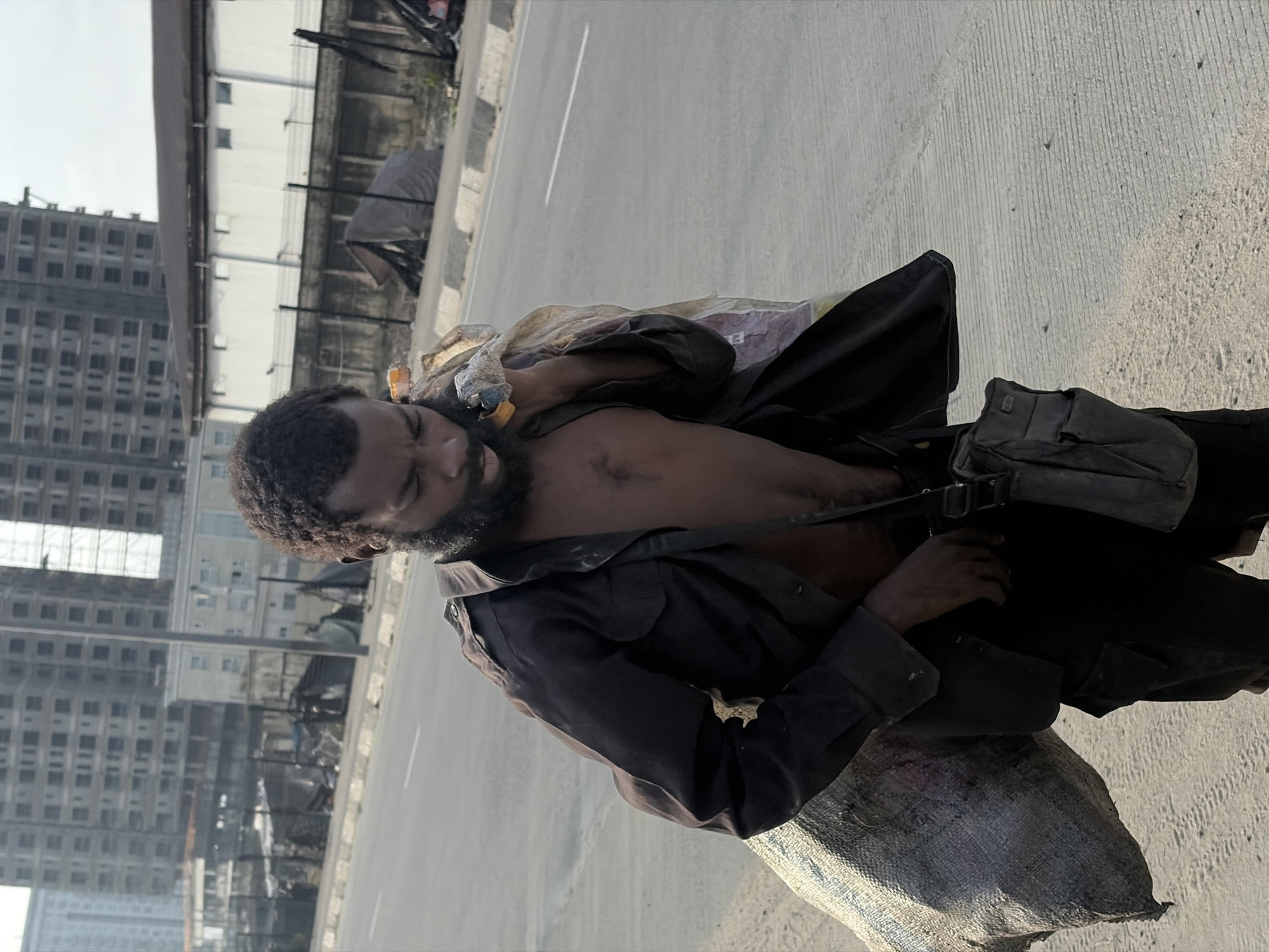 A Black man with dark skin, a beard, and curly hair walks along a paved roadside in Alaguntan, Nigeria. He is carrying a large, light-colored woven sack over his left shoulder and holding another plastic bag in his right hand. He wears a dark brown, open, collared shirt, revealing his chest and a dark undergarment. A dark bag is slung across his chest. In the background, there are multi-story buildings under construction, some with exposed framework, and a white building with a dark roof. A chain-link fence separates the road from the construction area. The sky is overcast.