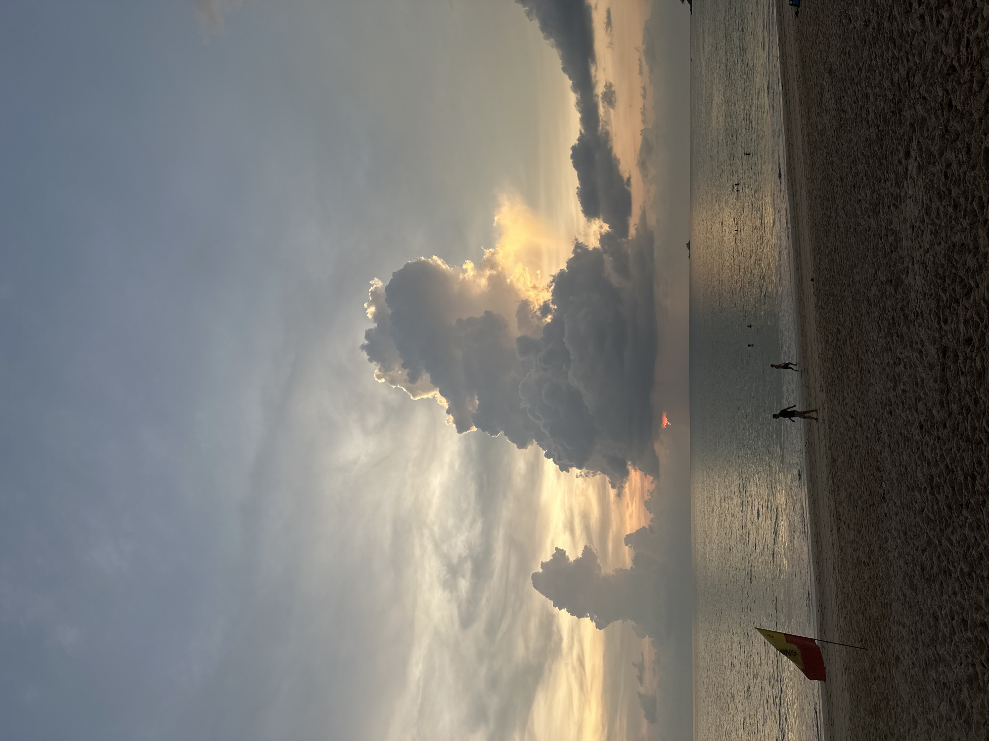 The image shows a beach scene at sunset in Choeng Thale, Thailand. The sky is filled with clouds, some dark and grey, others illuminated by the setting sun, creating a golden glow. The beach is sandy, and the ocean reflects the colors of the sky. There are a few people visible on the beach and in the water. A red and yellow flag is planted in the sand near the shoreline.