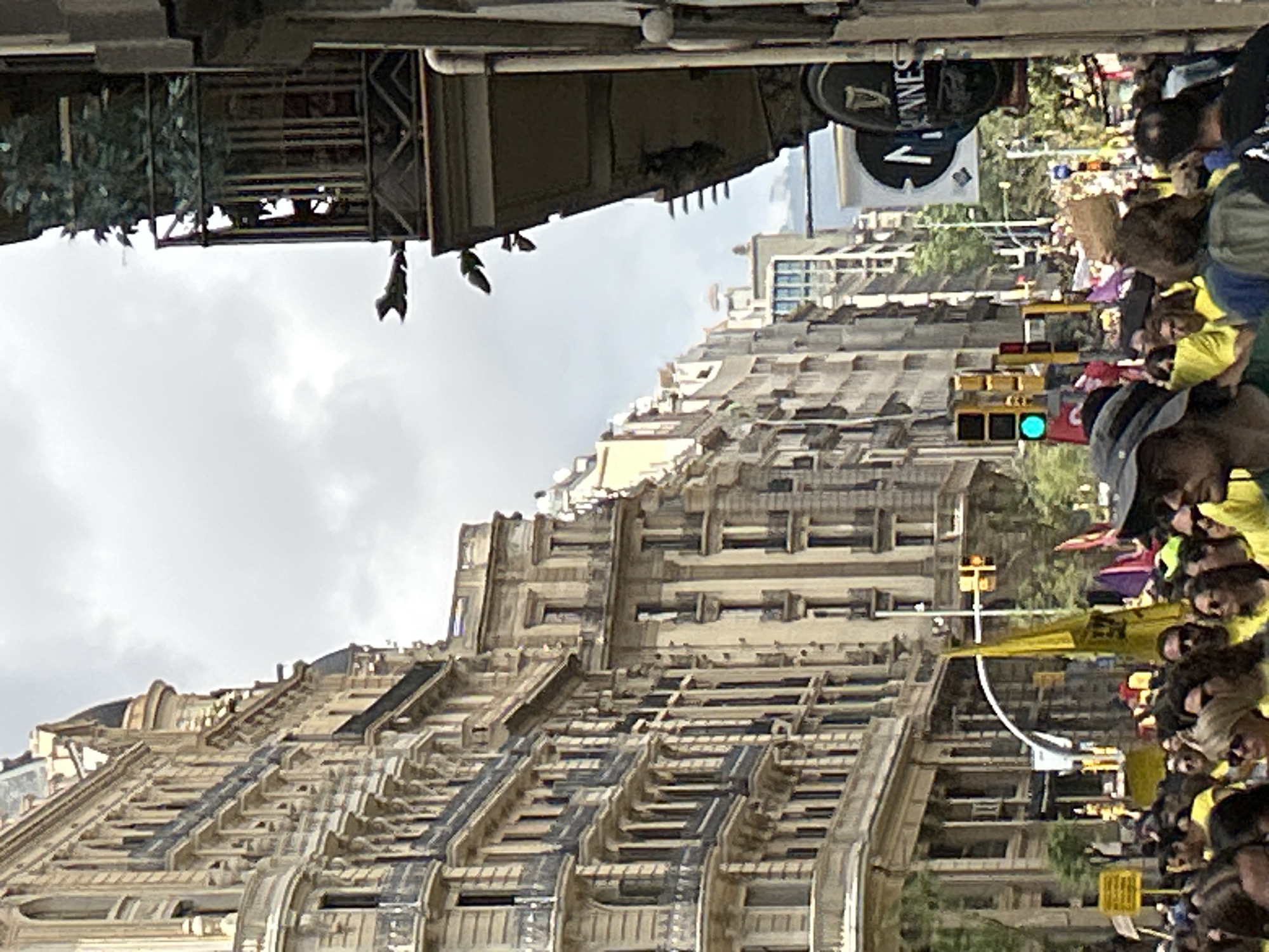 The image shows a street scene in Barcelona, Spain. The perspective is rotated 90 degrees clockwise. A crowd of people, many wearing yellow shirts, is walking down the street. A Guinness sign is visible in the background. The architectural detail of the building is ornate with dark and light sections. A traffic light is visible with a green light displayed. The sky is cloudy.