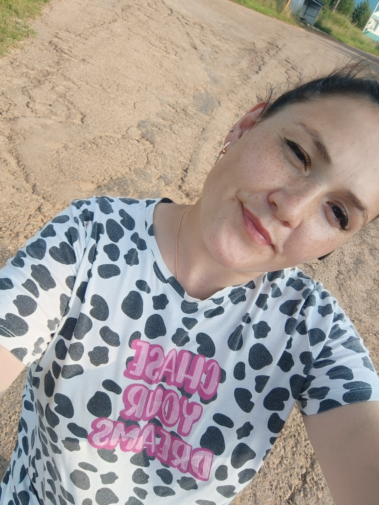 A woman is taking a selfie outdoors on a dirt road. She is wearing a white t-shirt with a black cow-print pattern and pink lettering that reads "CHASE YOUR DREAMS". The woman has fair skin, freckles, and dark hair pulled back. She is smiling slightly and looking at the camera. The background shows a dirt road with some grass and trees on the side. There is also a small building in the distance. The lighting suggests it is daytime. The location is the city of Karsha, Russia.