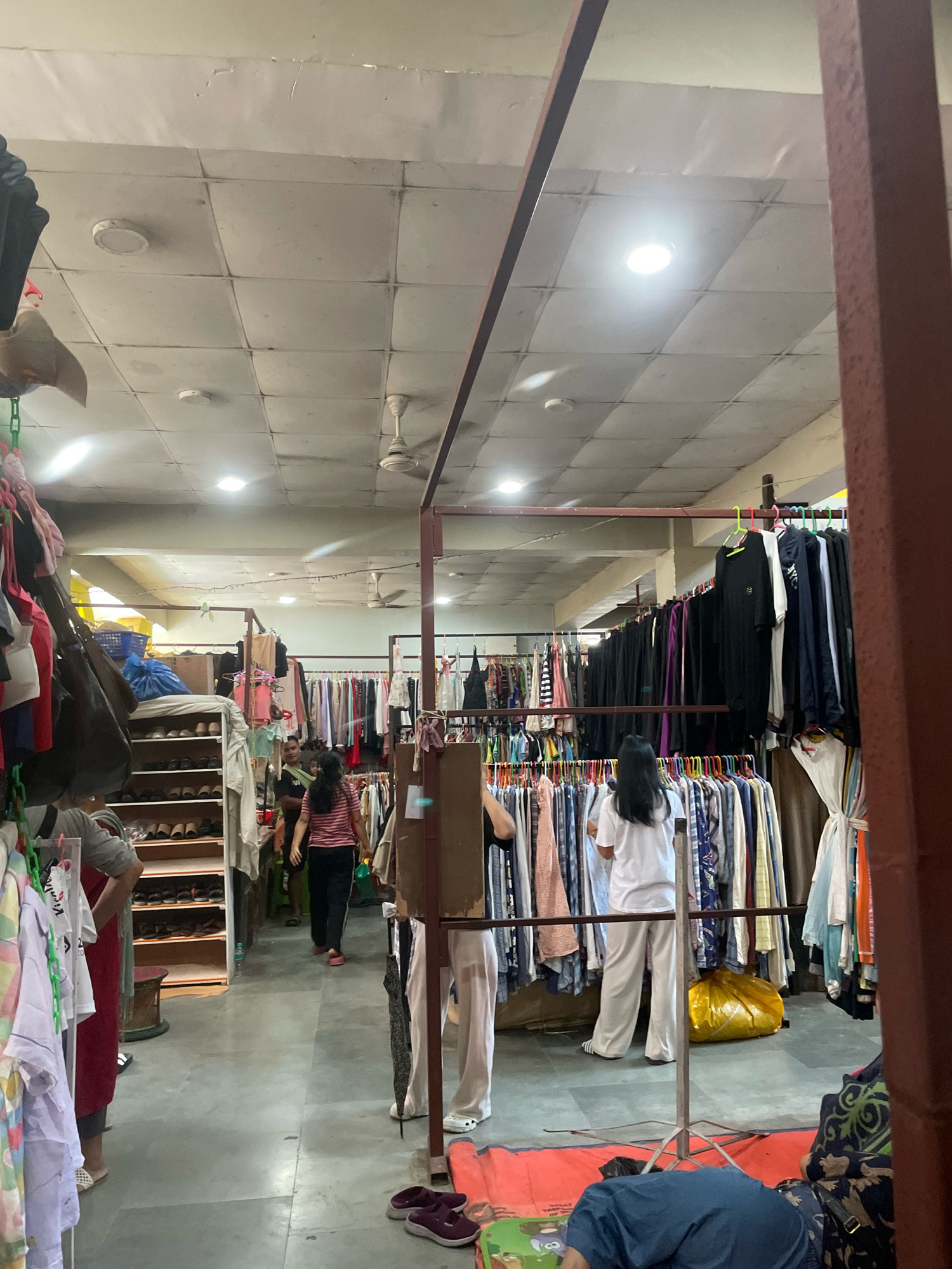 In Kangpokpi, India, an indoor marketplace is depicted with racks of clothing displayed for sale. Several individuals are present, including two women browsing through garments on racks. One woman, wearing a pink and black striped shirt and black pants, walks through the aisle. Another woman, dressed in a white t-shirt and off-white pants, stands in front of a clothing rack. A person, partially visible, is seen lying on the floor in the foreground. The environment features a tiled ceiling with fluorescent lighting and ceiling fans, indicative of a functional, albeit perhaps utilitarian, retail space. Racks of clothes, shoes on shelves, and bags are prominent objects within the scene.