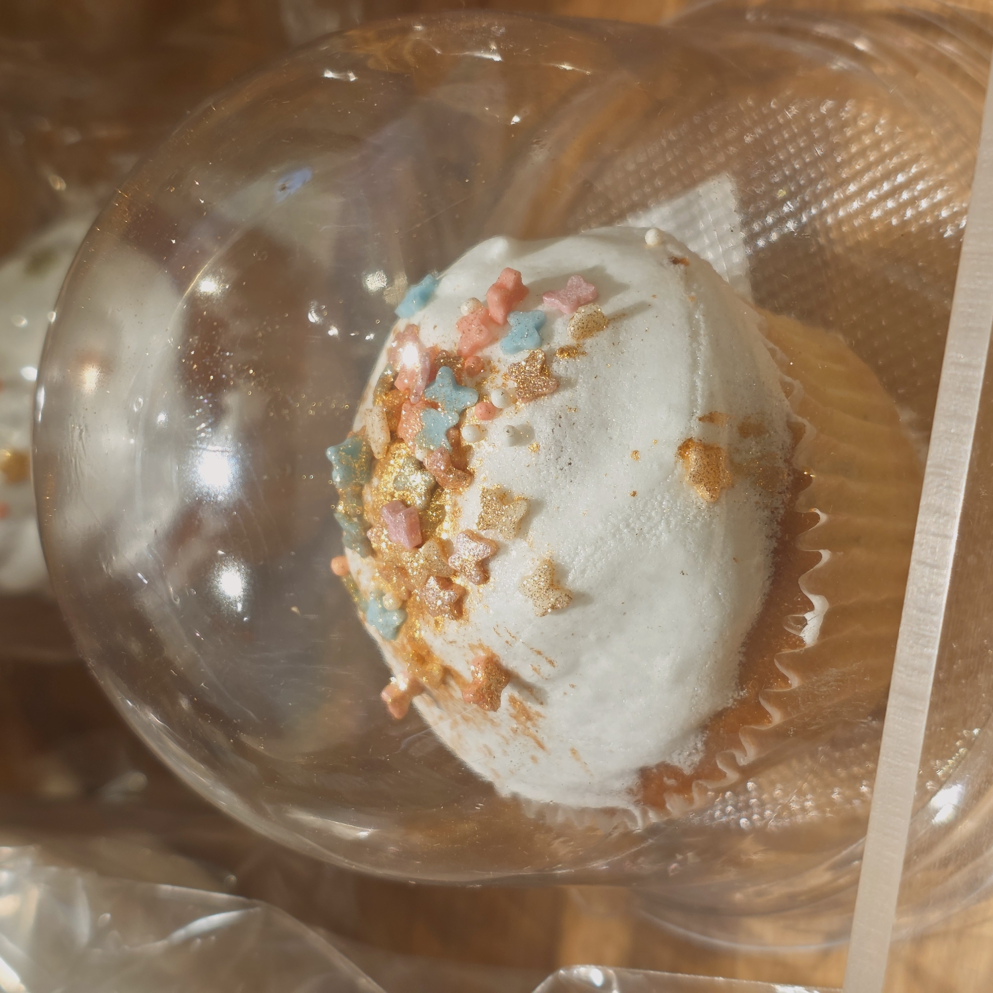 The image shows a cupcake in a clear plastic container. The cupcake has white frosting decorated with gold and pastel-colored star-shaped sprinkles. The background is slightly out of focus and appears to be a wooden surface. The image was taken in Saint Petersburg, Russia.