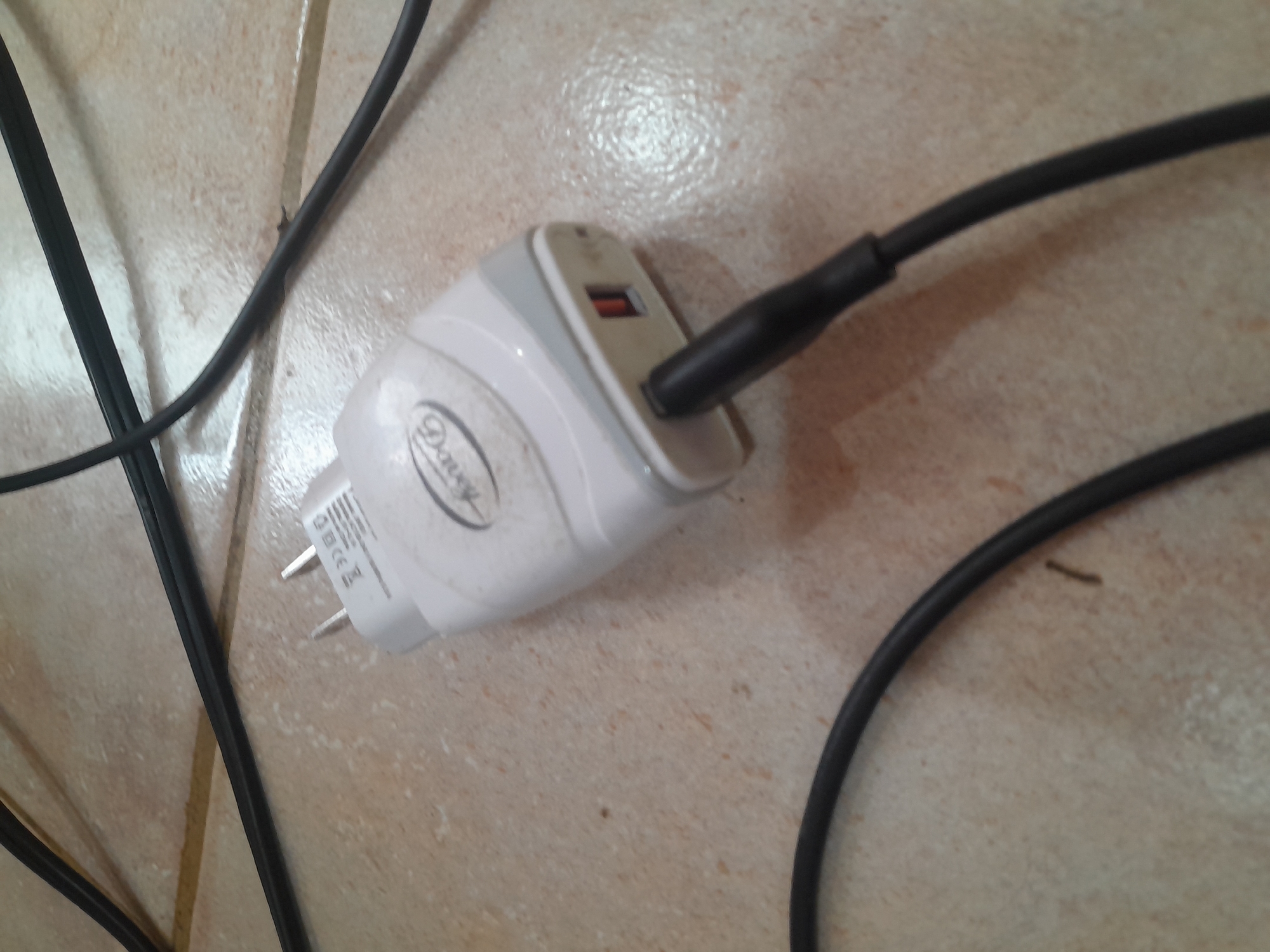 The image shows a white and gray USB charger plugged into a black cable, lying on a tiled floor. The charger has a logo that appears to say "Dauay" and some certification markings. The USB port is visible, as is the prongs for plugging into an electrical outlet. Black cables are also present, some running across the floor and out of frame. The environment appears to be an indoor setting, likely a residential or office space, based on the tiling. The location is identified as Santa Ana, Venezuela. There are no people or animals visible in the image. The lighting suggests it might be daytime, but the light source is not directly visible.
