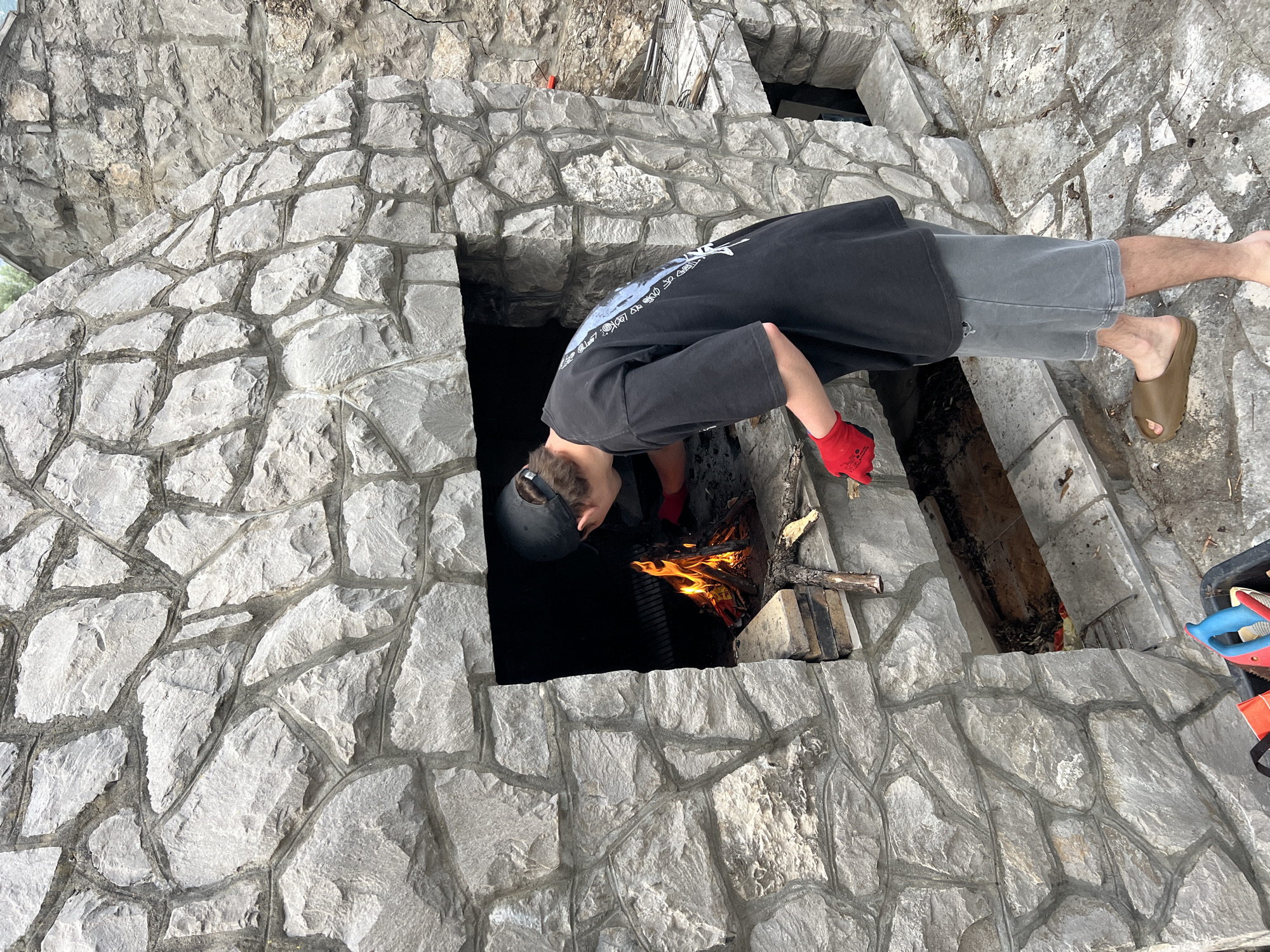 A person is leaning over a stone structure to tend a fire. The person is wearing a dark t-shirt, gray shorts, red gloves, a baseball cap, and brown sandals. The fire is burning inside a dark opening in the stone structure, which appears to be built into a wall or hillside. The stone structure is made of irregular gray stones. Part of the city of Ivanovići, Montenegro may be inferred from the surrounding elements.