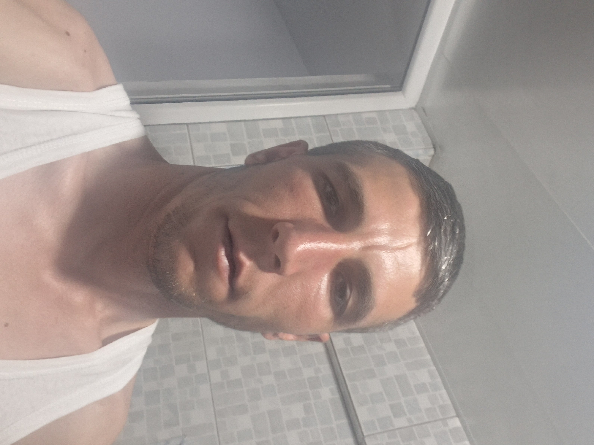 The image is a selfie of a light-skinned man in a white tank top, taken from the chest up. He has short, dark hair, a prominent nose, and a slight stubble. The background appears to be a bathroom, with tiled walls and a shower door visible. The lighting is bright, suggesting an indoor setting. The image does not contain any information about the city of Hovtamej, Armenia.