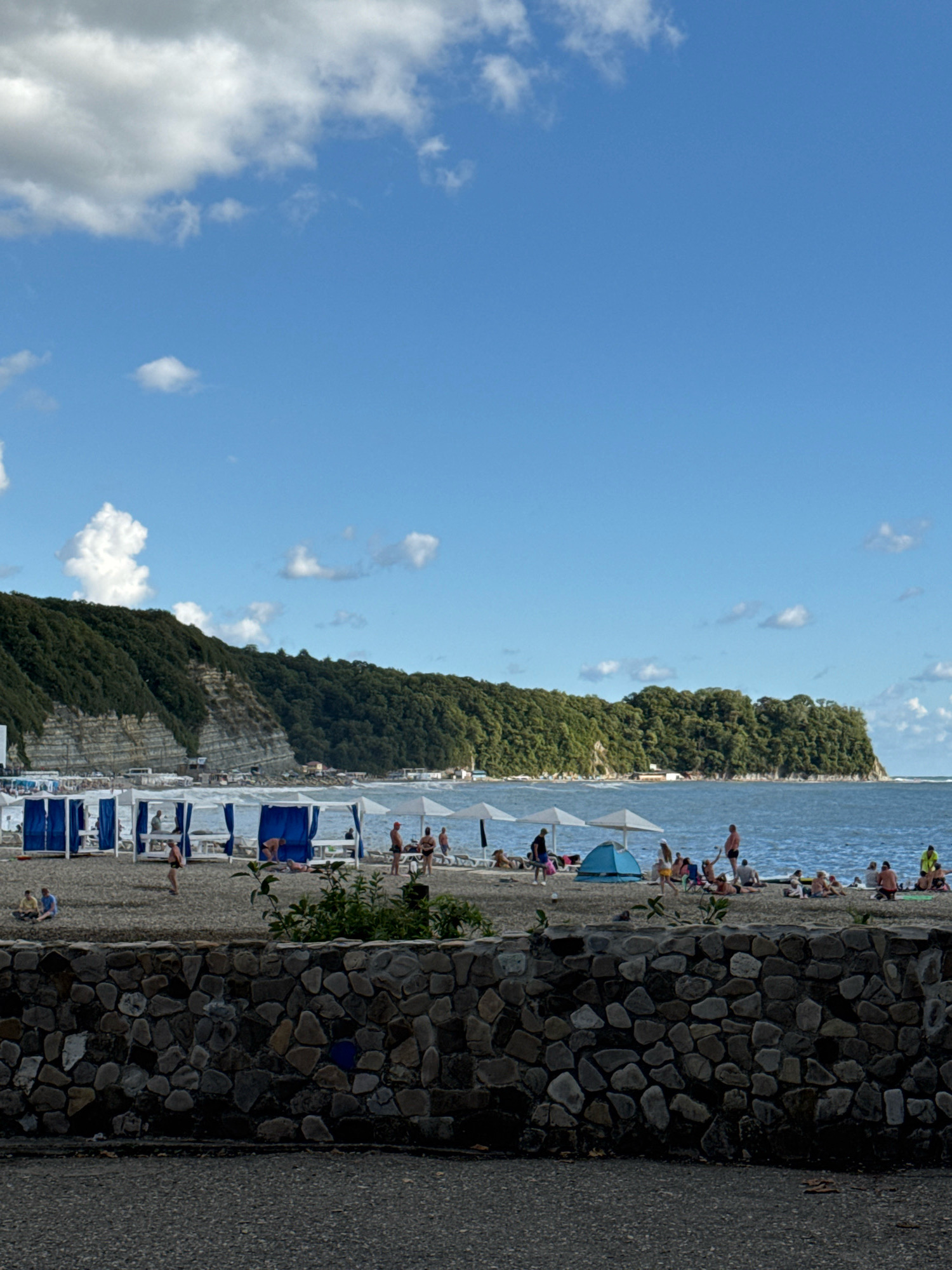 This is an eye-level, medium shot of a crowded beach in Novomikhaylovsky, Russia, under a bright blue sky with scattered white clouds. The foreground features a low, rough-hewn stone wall. Beyond the wall is a pebble beach with numerous blue and white canopies set up for shade. People are visible under these canopies and scattered across the beach, some sitting or lying down, while others are standing and interacting. The sea, a moderate blue, stretches across the mid-ground. In the background, a steep, densely forested hillside rises dramatically from the coastline. The overall scene depicts a popular summer recreational area.