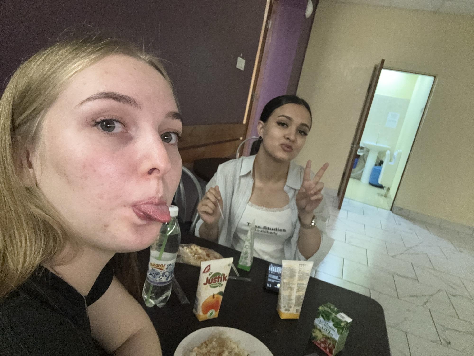 Two young women are in an indoor setting, possibly an apartment. The first woman, with blonde hair, is in the foreground, sticking her tongue out. The second woman, with dark hair pulled back, is seated further back and gives a peace sign. A table between them has several items on it, including a bottle of water, juice boxes, a plate of food, and other containers. In the background, there is a doorway leading to a bathroom. The location is indicated by the juice brand "Justik", which is distributed in Russia. Given the style and decor of the interior, it's plausible that this photo was taken in Perm, Russia.