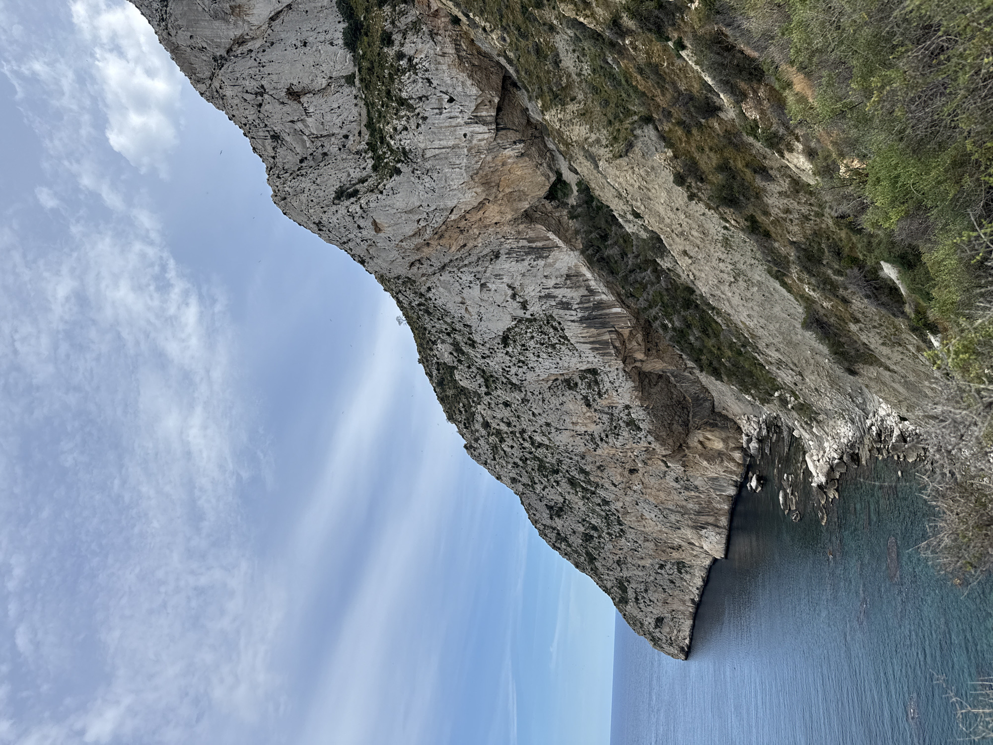 The image captures the Peñón de Ifach, a massive limestone rock formation rising from the Mediterranean Sea in Calp, Spain. The rock face is rugged and textured, with varying shades of gray and beige, speckled with patches of vegetation. The rock meets the water's edge, which is a mix of deep blue and turquoise hues. A clear blue sky with some thin, wispy clouds completes the scene.