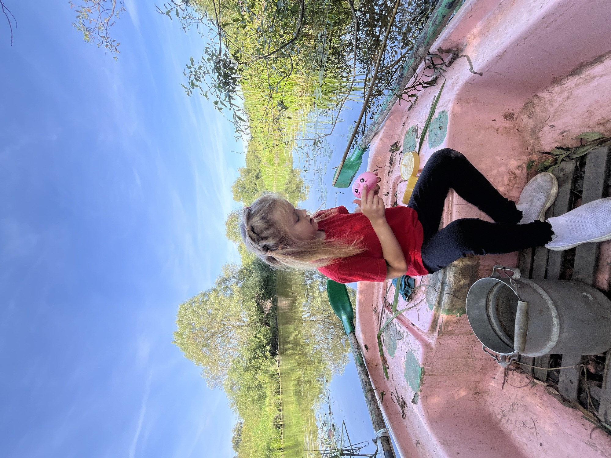 A young Caucasian girl with blonde hair in braids, wearing a red t-shirt and black pants, sits in a pink rowboat on a calm body of water, holding a pink toy. The boat is equipped with green oars. To the girl's left, an aluminum bucket rests on the wooden floor of the boat. The surrounding environment is lush with trees and reeds, reflecting in the water under a bright blue sky with scattered clouds. The location is identified as Sharkawshchyna, Belarus.