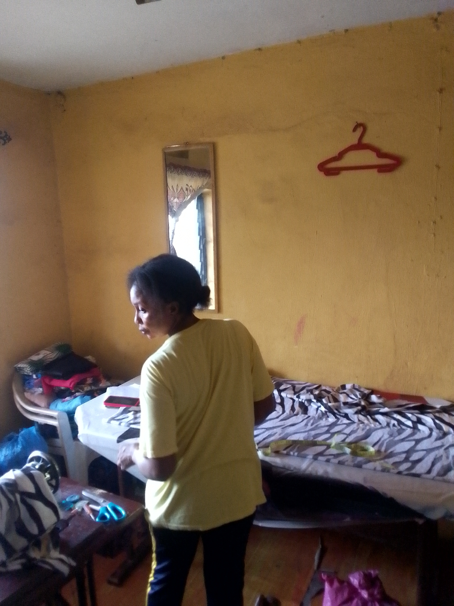 The image shows a person standing in a room in Unknown, Unknown. The walls are painted yellow and show signs of wear. There is a mirror on the wall, along with a red plastic hanger. A bed with a zebra-striped blanket is positioned against the wall. There's also a small table with clothes and other items on it. The person is wearing a yellow shirt and dark pants. A small wooden table with scissors is present near the bed.
