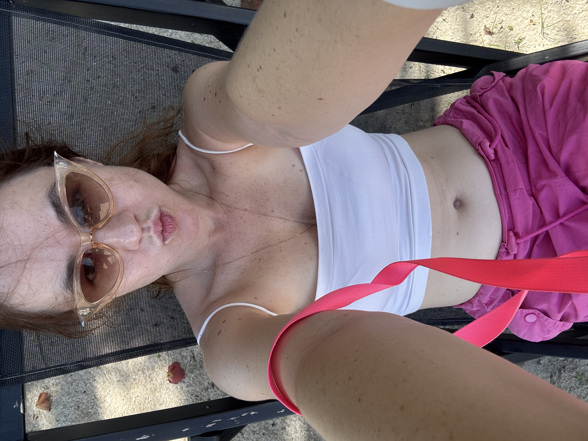 A woman is lying on a lounge chair outdoors, taking a selfie. She is wearing a white crop top and pink shorts. She has on large, light-colored sunglasses and her hair is down. A pink strap is visible across her body. The background shows a textured surface that appears to be sand or dirt with some scattered leaves. The lighting suggests it is daytime. The scene is casual and relaxed, possibly a backyard or poolside setting.