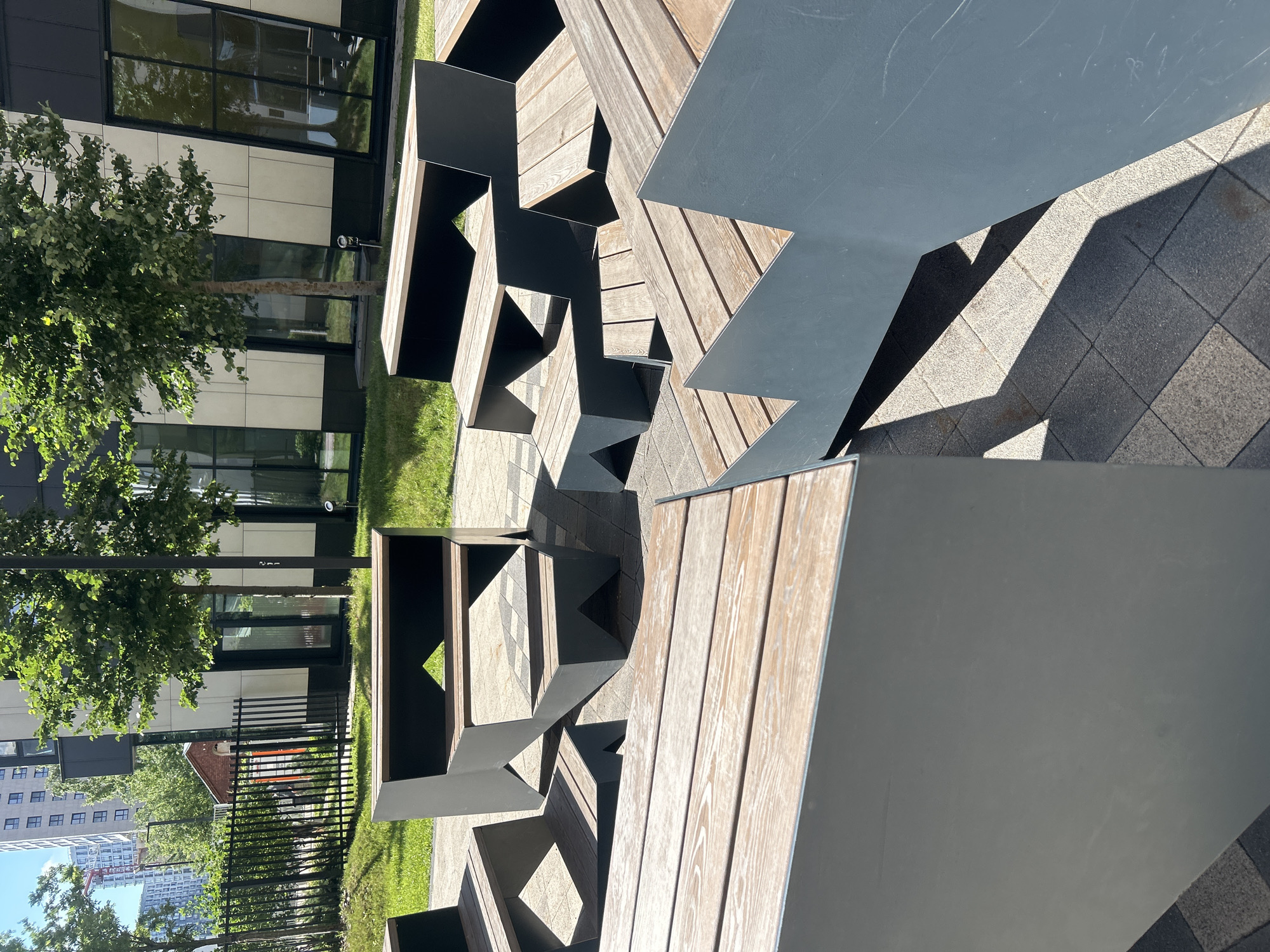 The image shows a modern outdoor seating area composed of tiered, angular benches with wooden slats and gray metal frames. The benches are arranged in a cluster on a paved area with a visible pattern of square tiles. Green grass and trees are visible behind the seating area, and in the background, there are tall apartment buildings, suggesting an urban environment. One of the buildings in the background has a crane in front of it, indicating ongoing construction. The lighting suggests it is daytime, with distinct shadows cast by the benches onto the paving stones. The location is identified as Moscow, Russia. There are no people present in the image.