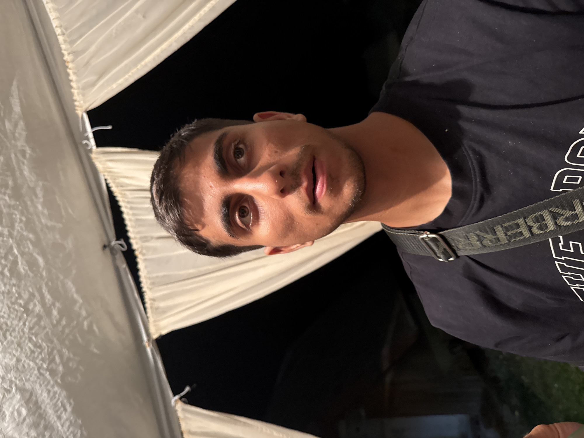 A head and shoulders, medium close-up shot shows a young man with short, dark hair, dark eyes, and a light complexion. He is wearing a dark blue t-shirt with a thick strap crossing his chest from his left shoulder to his right. The strap has a repeating pattern of letters that appear to be "BER." The man is looking directly at the camera with a neutral expression. Behind him, white fabric drapes and billows, creating a tent-like structure. The background is dark, suggesting it is nighttime. The environment appears to be outdoors, possibly at an event or gathering in the city of Sredets, Bulgaria.