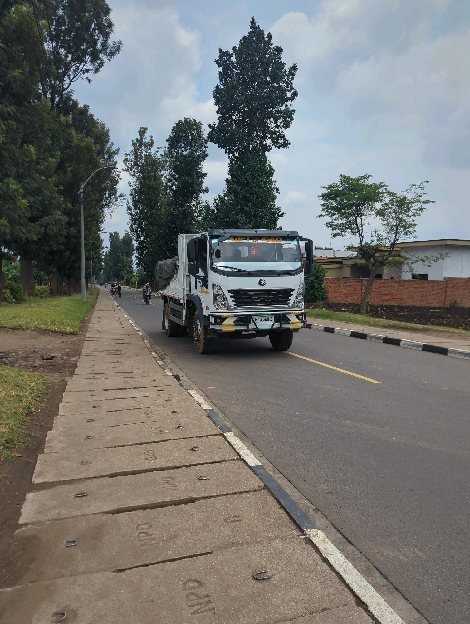A white truck with a covered bed is driving on a road in Musanze District, Rwanda. The truck has a license plate that reads "RAJ 308 J" and a message on the windshield that appears to be "GLORY OF GOD". The road has a yellow dividing line and a black and white striped curb. To the left of the road is a sidewalk made of concrete slabs. Beyond the sidewalk, there are trees and grass. In the background, there are buildings and more trees. Several people are visible in the distance, including someone riding a motorcycle and people in what appear to be carts being pulled by animals. The sky is cloudy.