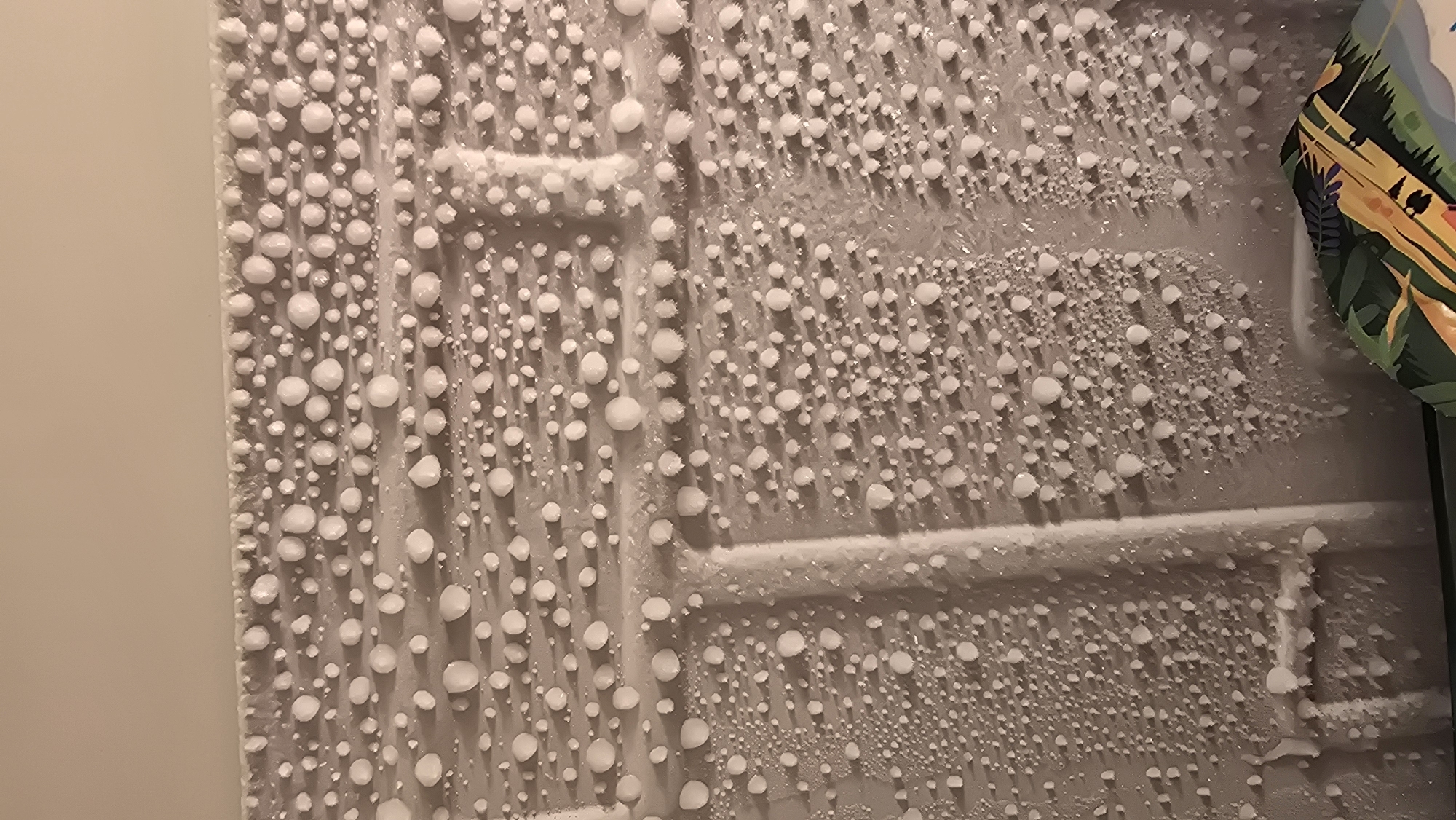 This image depicts a close-up, overhead view of hailstones accumulated on a grey, textured surface, possibly a wall or a patterned mat. The hailstones are small and spherical, varying in size from tiny pebbles to marbles. They are densely packed, particularly along horizontal lines, creating patterns of accumulation. Some of the hailstones appear to be melting or have a wet surface, indicated by slight discoloration and a sheen. The background is a plain, light-colored wall or ceiling. The overall impression is of a recent hailstorm. The location is specified as Novohuivynske, Ukraine, suggesting this is an outdoor scene where hail has fallen.