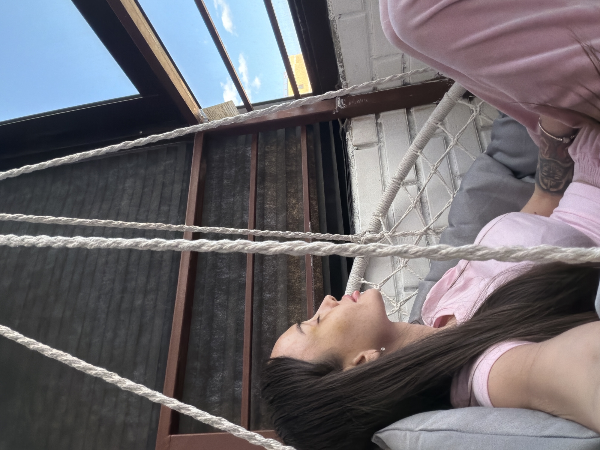 A woman lies in a hammock indoors. Her head rests on a gray pillow. She wears a pink shirt. A tattoo is visible on her left arm. The hammock is suspended from a dark brown metal frame. White brick walls are visible to the right of the frame. Gray blinds are visible behind the frame. Above the frame, a partial view of the sky is visible through a window. Based on architectural characteristics, the location is likely in Saint Petersburg, Russia.