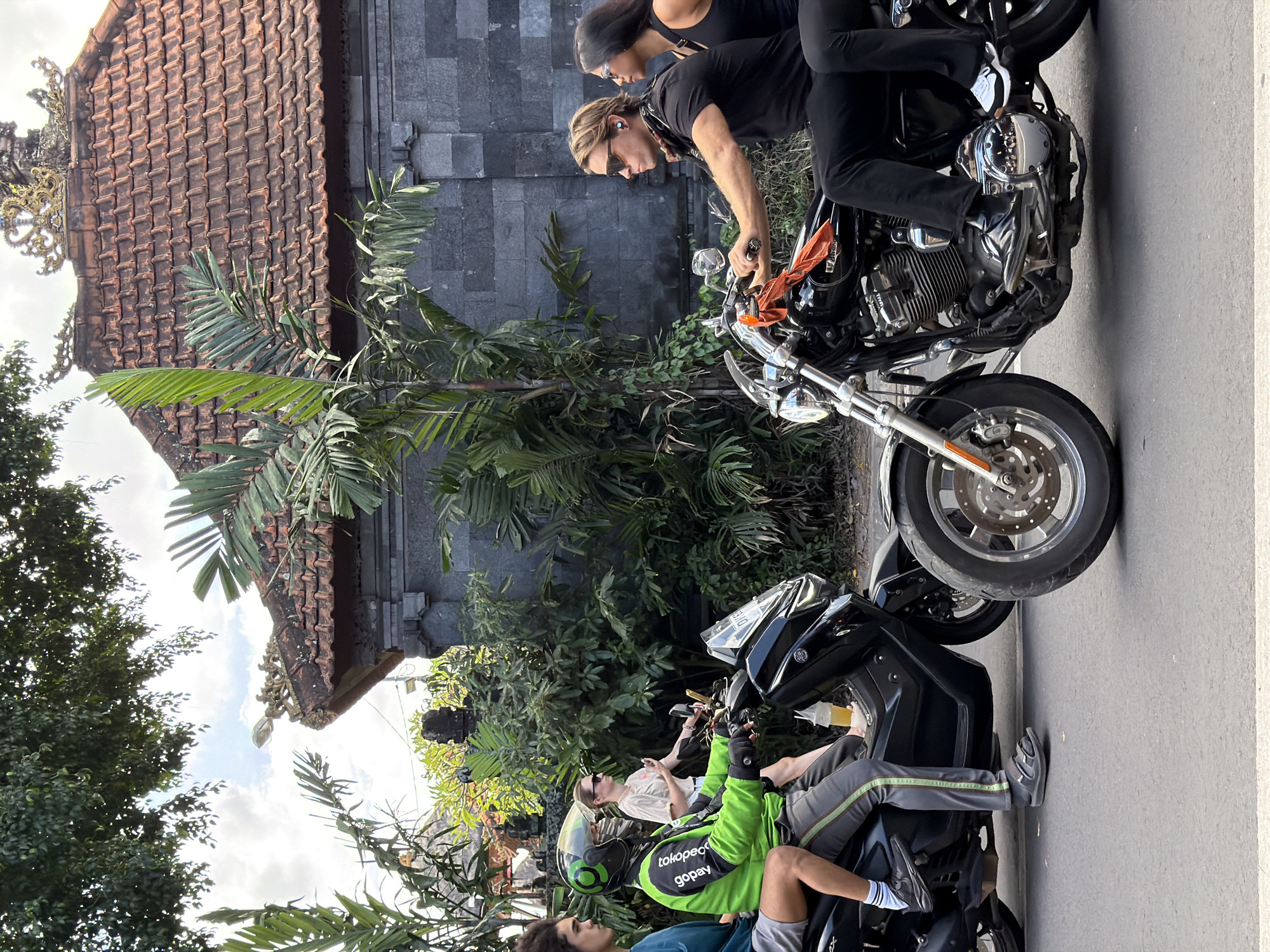 This media file depicts three people on motorcycles in North Kuta, Indonesia. The scene is outdoors, with a building featuring a tiled roof and stone walls in the background. Lush green foliage, including palm fronds, is abundant, suggesting a tropical environment.
In the foreground, two motorcycles are parked side-by-side. The person on the front motorcycle is wearing a bright green jacket with the text "tokopedia gopay" visible. They also have a helmet on. The person on the second motorcycle is wearing a white t-shirt and sunglasses. Behind them, two people are on another motorcycle. The person in front is wearing a black tank top and sunglasses, while the person in the back is wearing a black t-shirt and black pants. An orange cloth is tied to the handlebars of this motorcycle.
The time of day appears to be daytime, possibly late morning or early afternoon, given the lighting conditions and the presence of shadows. The weather seems to be warm and pleasant. The overall mood is casual, suggesting a leisurely ride or a stop during a journey. The architecture and vegetation strongly indicate a location in Southeast Asia, consistent with the provided location context of North Kuta, Indonesia.