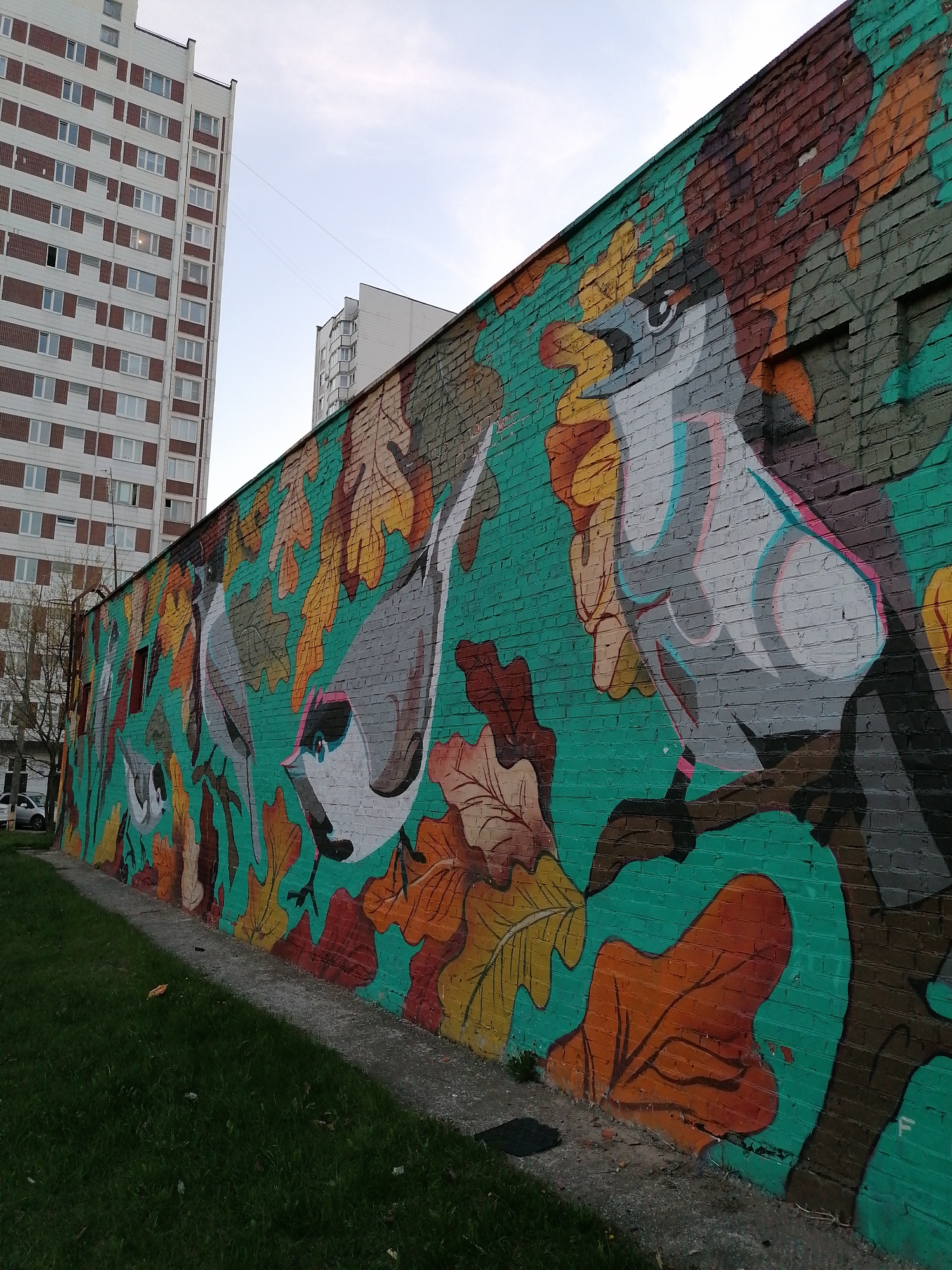The image shows a long, brick wall with a vibrant mural of birds and autumn leaves. The wall is painted in shades of green, brown, orange, and yellow. Several birds, depicted in a stylized manner with grey and white plumage, are perched among the leaves. The mural covers the entire length of the wall, which appears to be adjacent to a grassy area. In the background, tall apartment buildings with brick accents are visible against a pale sky. The location is Unknown, Unknown.