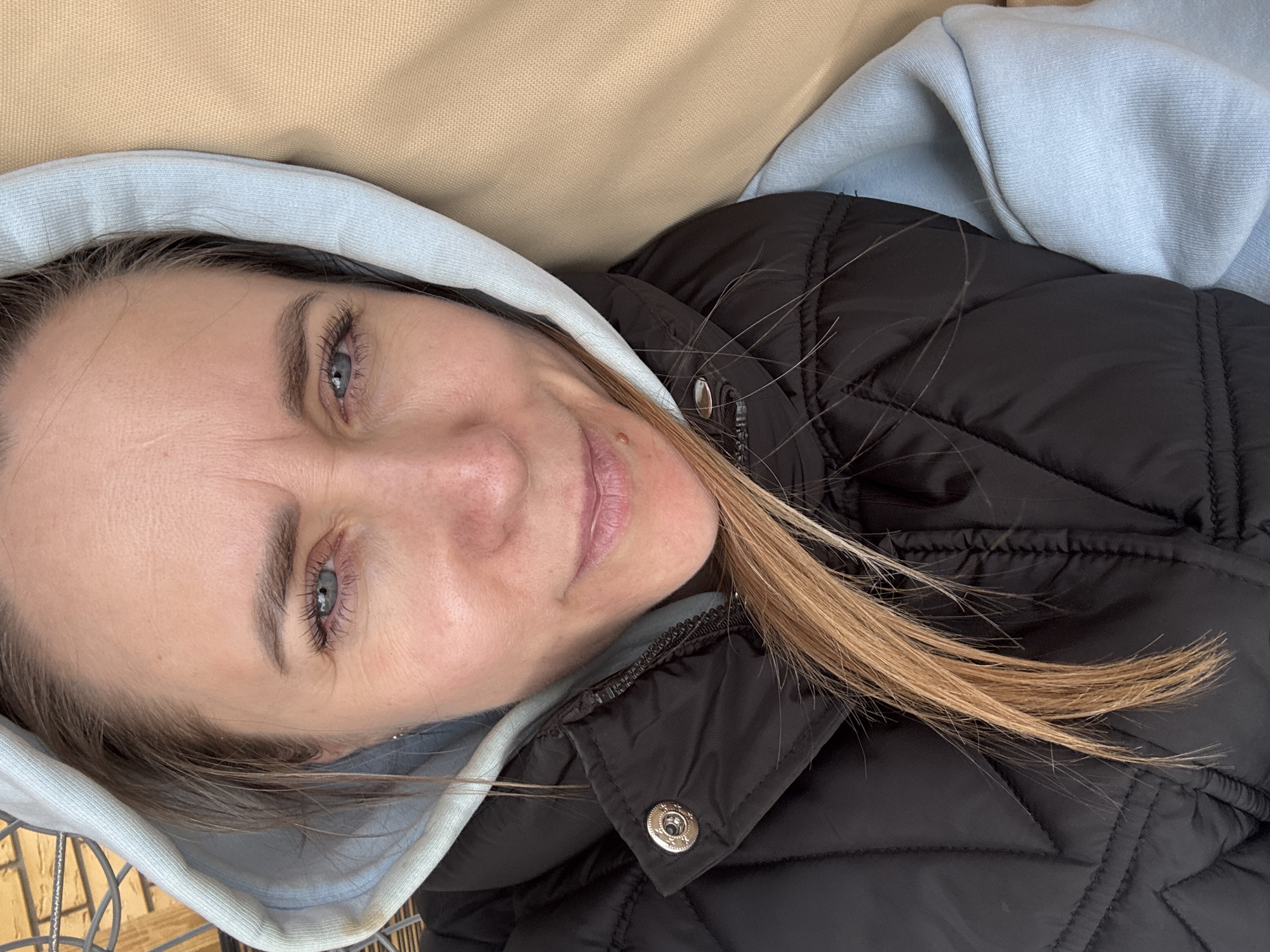 A woman with light skin and light blue eyes is lying down, possibly indoors. She is wearing a light blue hoodie with the hood up and a black puffy jacket. Some strands of blonde hair are visible over the jacket. The background is a blurred, beige fabric. The photo may be taken in Chaikovsky, Russia, though this cannot be verified from the image alone.
