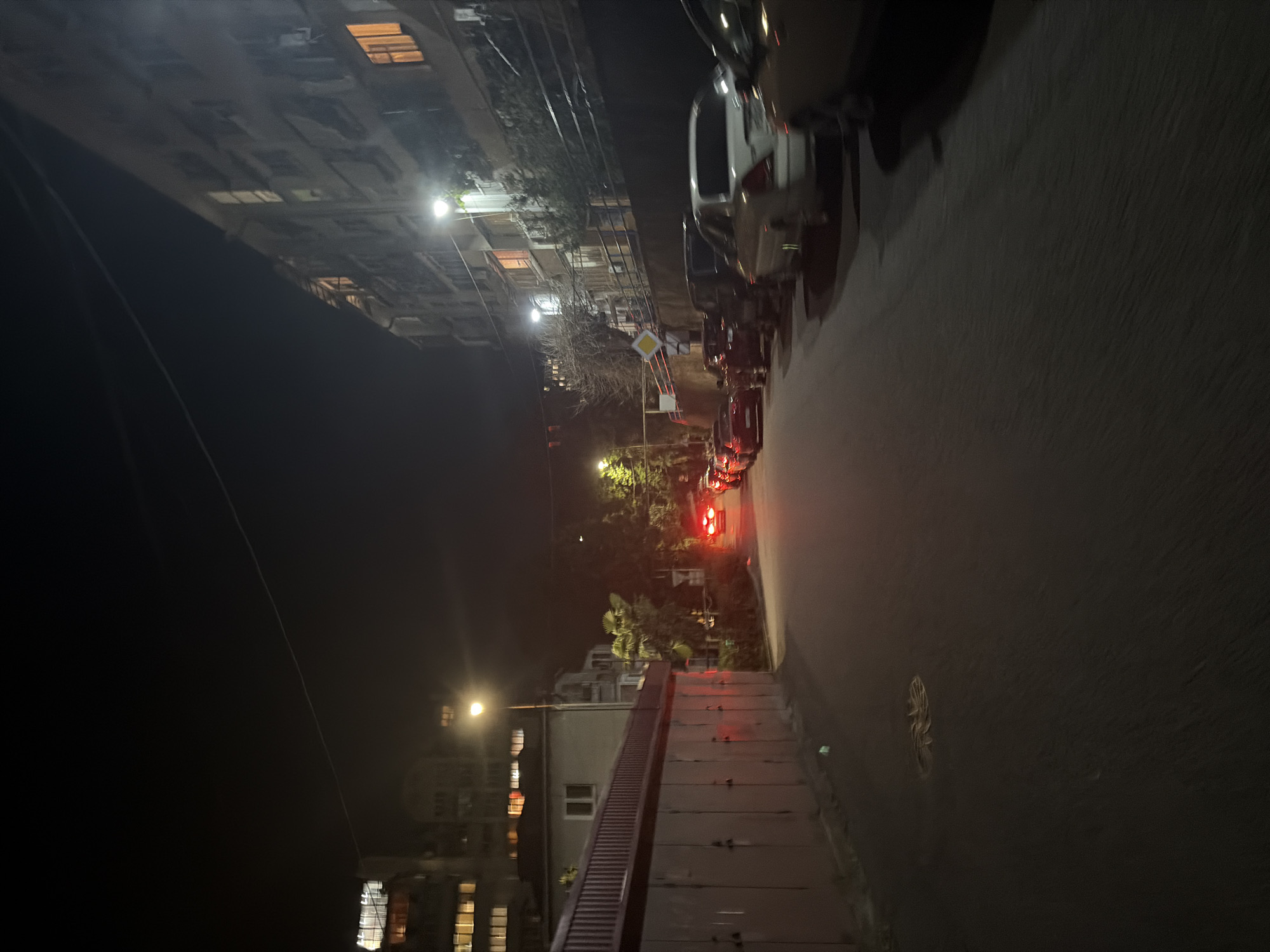 The image displays a nighttime urban street scene in Sochi, Russia, captured from a slightly elevated vantage point. The sky is dark, characteristic of late hours, with artificial light sources illuminating the environment.
On the left side of the frame, several multi-story buildings, likely residential or commercial, line the street. Many of their windows glow with warm, inviting light, suggesting occupancy. Numerous bright streetlights are scattered throughout the scene, casting circular pools of light that reflect on the ground and building facades. A thin, dark cable or power line stretches across the upper left portion of the dark sky.
The middle and right sections of the image feature a street or parking area filled with various parked cars, including sedans and larger vehicles. Some of these cars exhibit illuminated red taillights. Towards the back, several trees are visible, some with deciduous branches and others appearing to be palm trees, typical of Sochi's climate, interspersed among the buildings. A prominent yellow, diamond-shaped traffic sign, commonly indicating a priority road, stands on the right side of the street.
The immediate foreground is dominated by a wide, dark, and textured surface, possibly a flat roof or a concrete structure. On its right side, a sloped, ribbed structure, resembling a ramp or a set of wide stairs, descends into the midground.
The scene is devoid of visible people or activity, conveying a quiet and still atmosphere typical of an urban area at night. There is no discernible text on any signs, vehicles, or buildings.