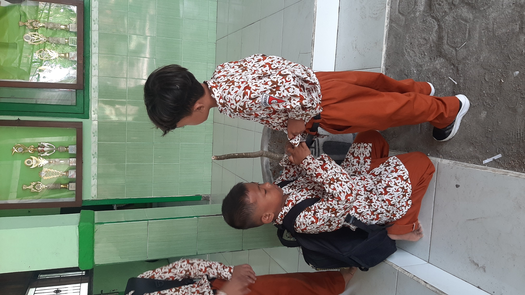 The image depicts two young boys wearing school uniforms in what appears to be a school hallway or common area in Sumberjo, Indonesia. Both boys have short dark hair. The boy on the left is kneeling and wearing a white shirt with a repetitive brown and red pattern and orange pants. He has a black backpack positioned behind him and appears to be adjusting something on his wrist. The boy on the right is standing and is also wearing a similar patterned shirt and orange pants. He is looking down and interacting with the kneeling boy. Behind the boys, the wall is tiled with light green square tiles, and there are display cases containing trophies mounted on the wall. A small tree in a pot is also visible in the background. The floor is a mixture of tiled and dirt surfaces.
