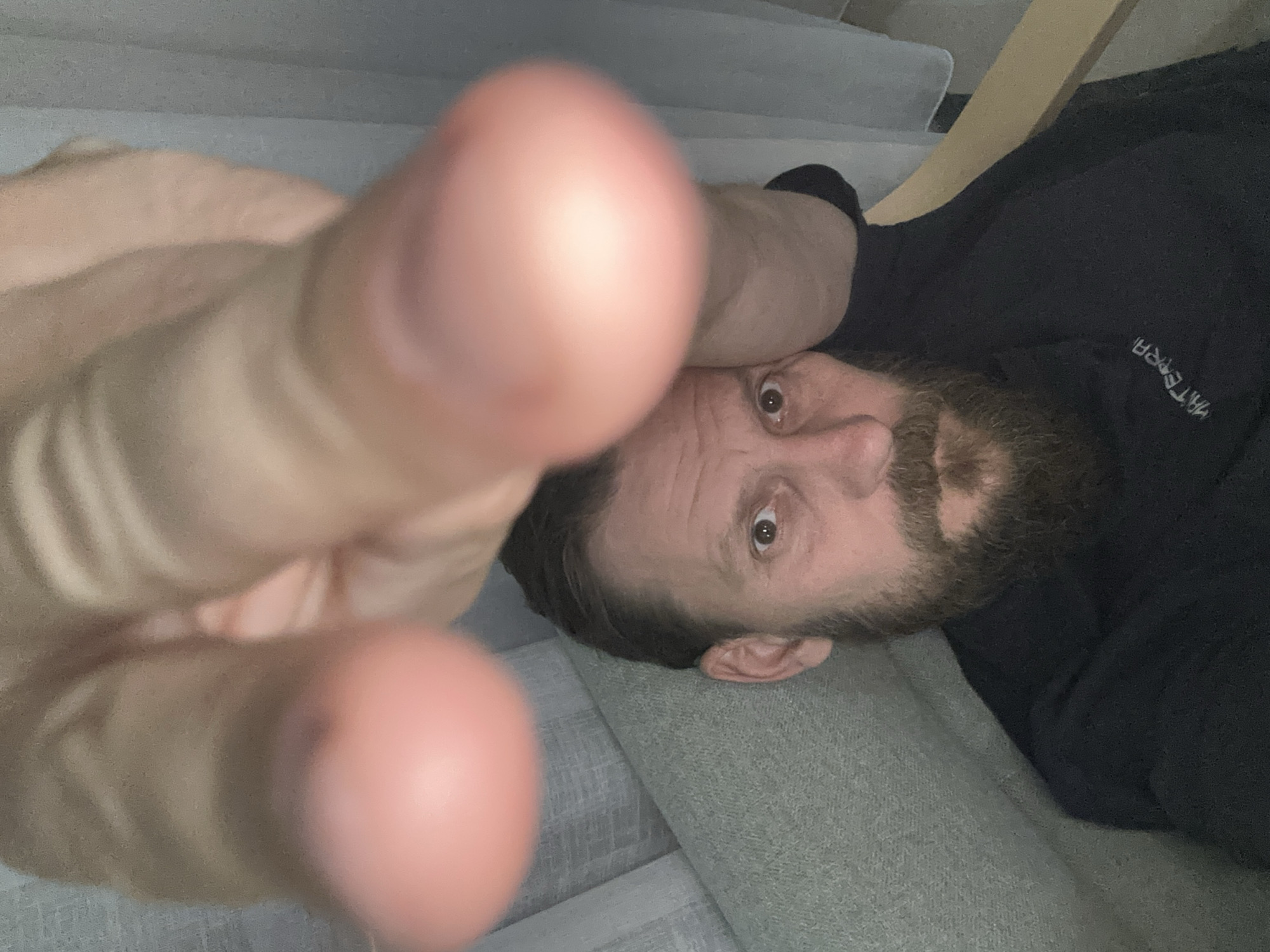 A selfie photograph of a man lying down, with his face visible at the lower right. His hand, with fingers extended towards the camera, is significantly out of focus and dominates the left side of the frame. The man has a beard and is wearing a dark-colored top with the text "MATERRA" visible. He appears to be reclining on a light grey upholstered surface. No landmarks or information are visible that definitively place this image in Fethiye, Turkey.