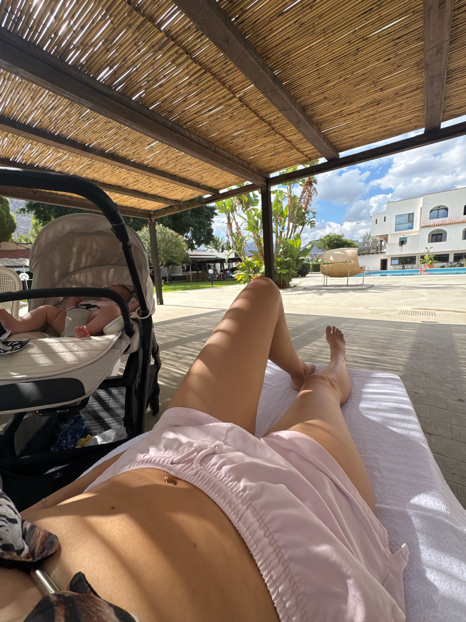 This image captures a serene poolside scene in Isola delle Femmine, Italy, under a shaded bamboo pergola. In the foreground, a person is relaxing on a lounge chair, their legs and torso visible, sun-kissed and clad in light pink shorts. Nearby, a baby rests peacefully in a stroller, its tiny feet peeking out from the soft bedding. Beyond the immediate lounging area, a clear blue swimming pool shimmers, bordered by lounge chairs and a modern white building with arched windows. Lush green foliage and trees frame the background, with a hint of mountains in the distance under a bright, partly cloudy sky. The overall atmosphere is one of tranquility and vacation relaxation.