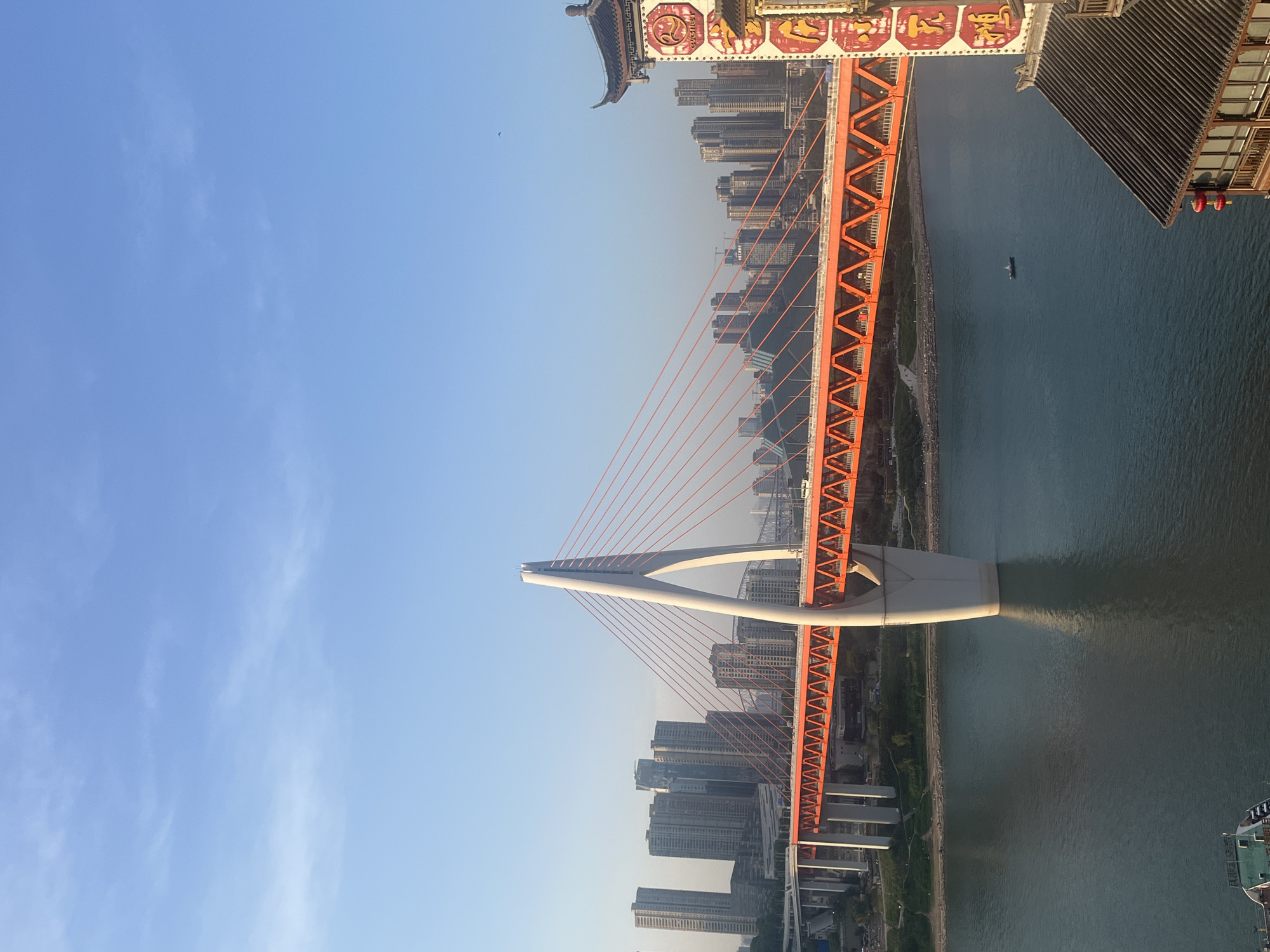 The image shows the Dongshuimen Bridge in the city of Yuzhong District, China. The bridge is orange and white and spans a river. The sky is blue. There are buildings on either side of the river. A pagoda is on the left of the upper part of the frame.