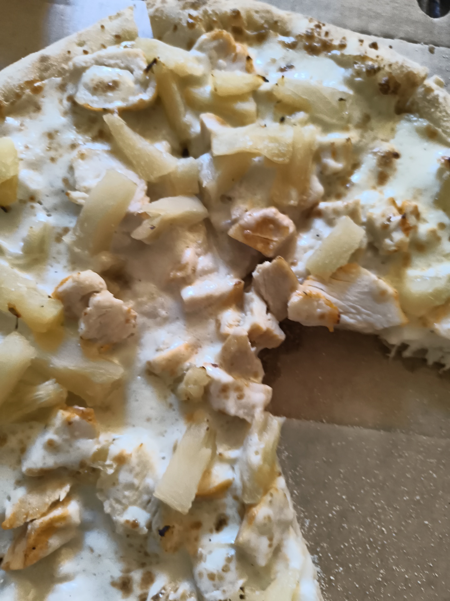 The image is a close-up, overhead shot of a pizza, likely a Hawaiian pizza with chicken and pineapple, in a cardboard pizza box. The pizza has a white sauce base, topped with chunks of cooked chicken, pieces of pineapple, and what appears to be melted mozzarella cheese. Some parts of the crust are visible, and the pizza box is partially visible in the background. The lighting suggests an indoor setting, possibly a restaurant or home. There are no people or specific landmarks visible in the image. The image quality is slightly blurry and oversaturated, making it difficult to discern fine details. It is not possible to determine the specific location within Mayma, Russia from this image.