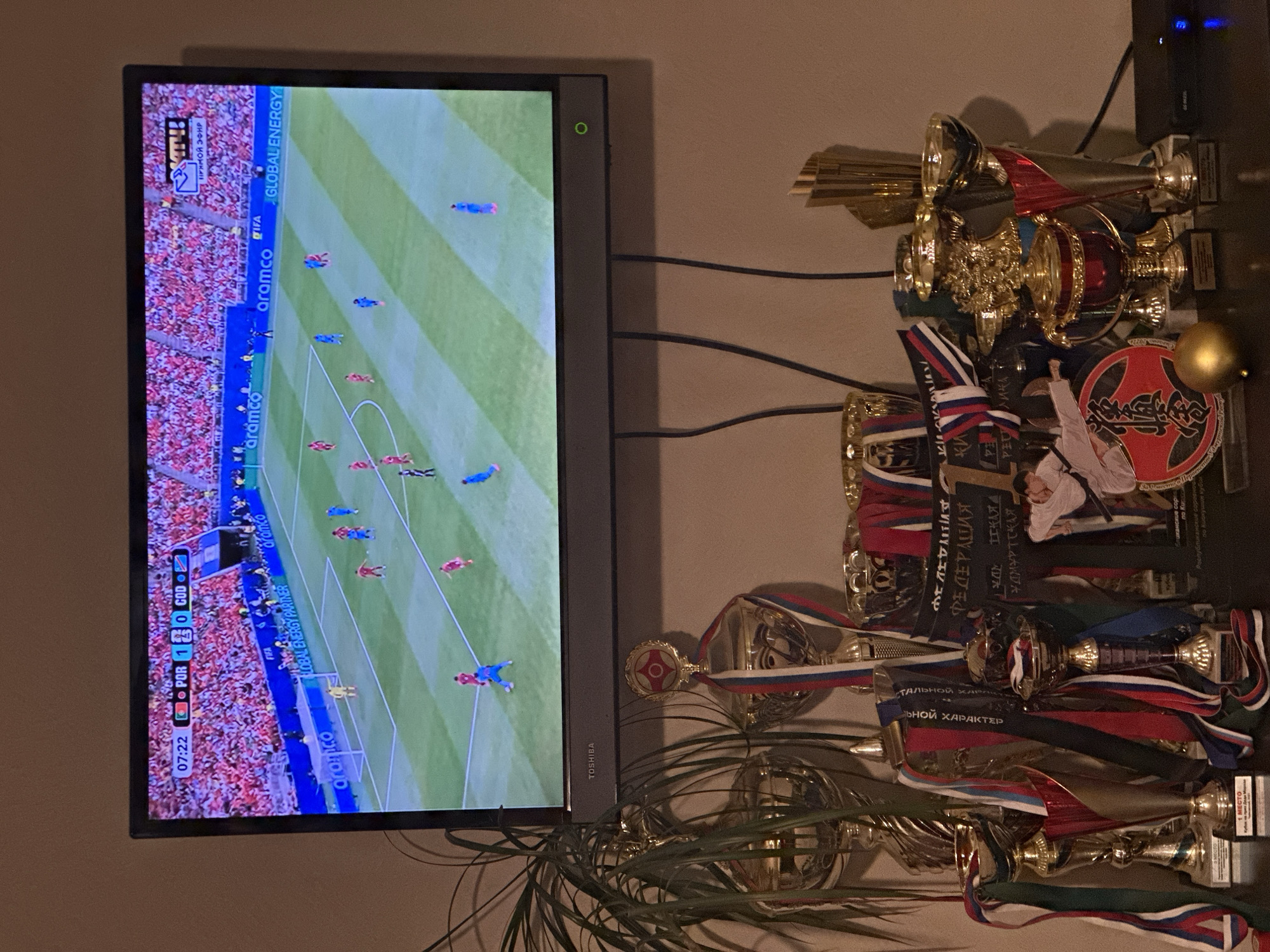 This media file captures a domestic scene featuring a television displaying a soccer match and a collection of trophies. The television screen shows a game in progress with a scoreboard indicating "POR 1" versus "COD 0" and a timestamp of "07:22". The stadium audience is visible in the background of the broadcast. Prominently displayed next to the television are several gold trophies of varying sizes, adorned with ribbons in red, white, and blue, suggesting a connection to Russian national colors. One of the trophies has a circular emblem with a red star and martial arts imagery. Text on the trophies and ribbons includes Russian inscriptions such as "ФЕДЕРАЦИЯ" (Federation), "УДМУРТСКАЯ" (Udmurtian), and "ТАЛЬНОЙ ХАРАКТЕР" (Total Character). The setting appears to be a living room or entertainment area, with cables visible behind the television and a plant in the foreground. The overall impression is one of sports enthusiasm and recognition of achievement within a home environment. The image is likely taken indoors, given the lighting and the presence of a television and trophies. There are no clear indicators of the time of day or weather conditions. The location context is Chaikovsky, Russia, which aligns with the Russian text visible on the trophies.
