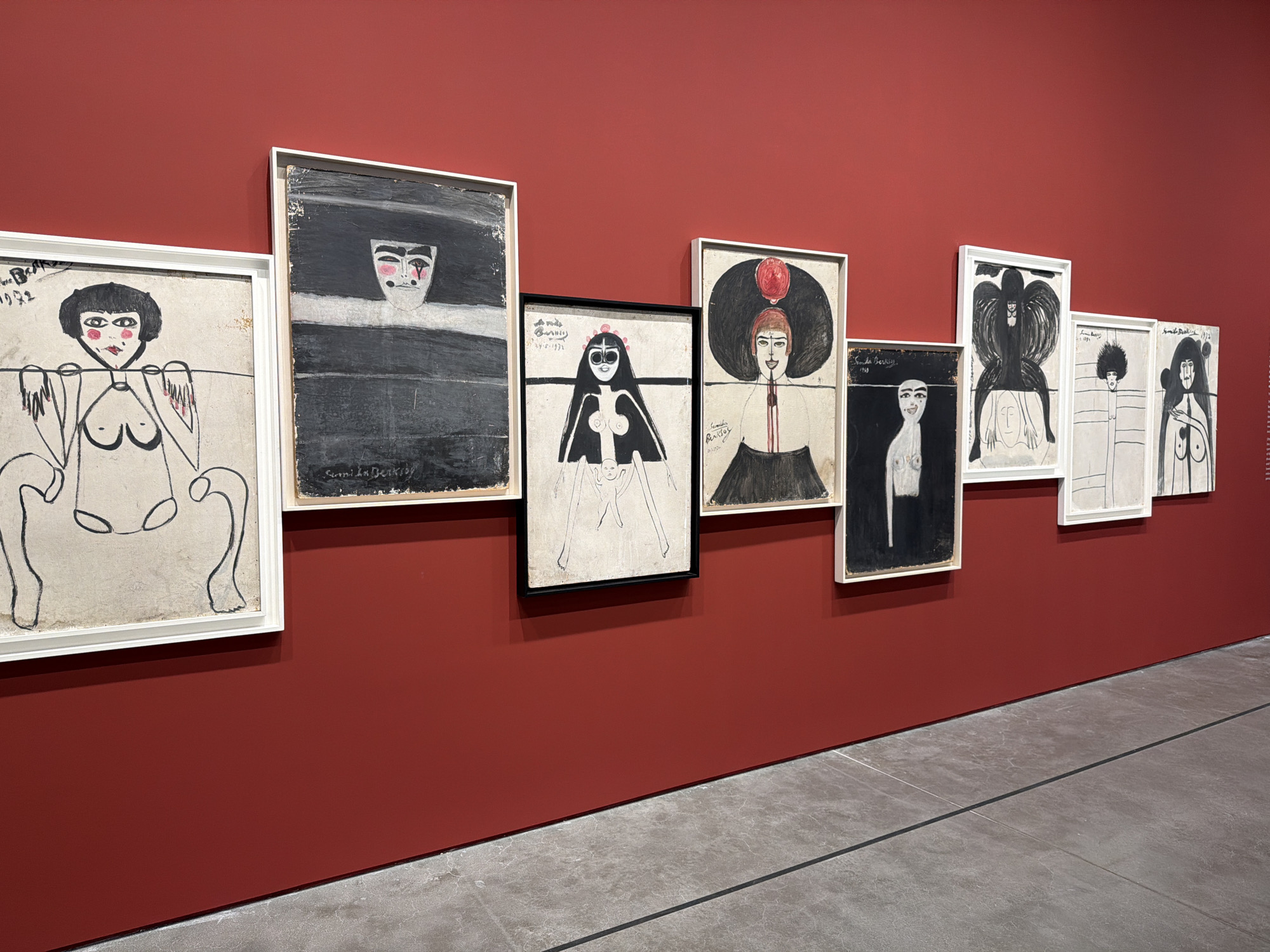 The image shows a series of seven framed artworks displayed along a red wall in a gallery or museum. The wall is a deep, solid red, and the floor is a gray, polished stone. The artworks appear to be paintings or drawings, primarily in black and white with some touches of red, featuring stylized depictions of human figures. The frames are mostly white, with one black frame. The artworks vary in size and orientation and are hung in a linear arrangement along the wall. The location may be in Beyoğlu, Turkey.