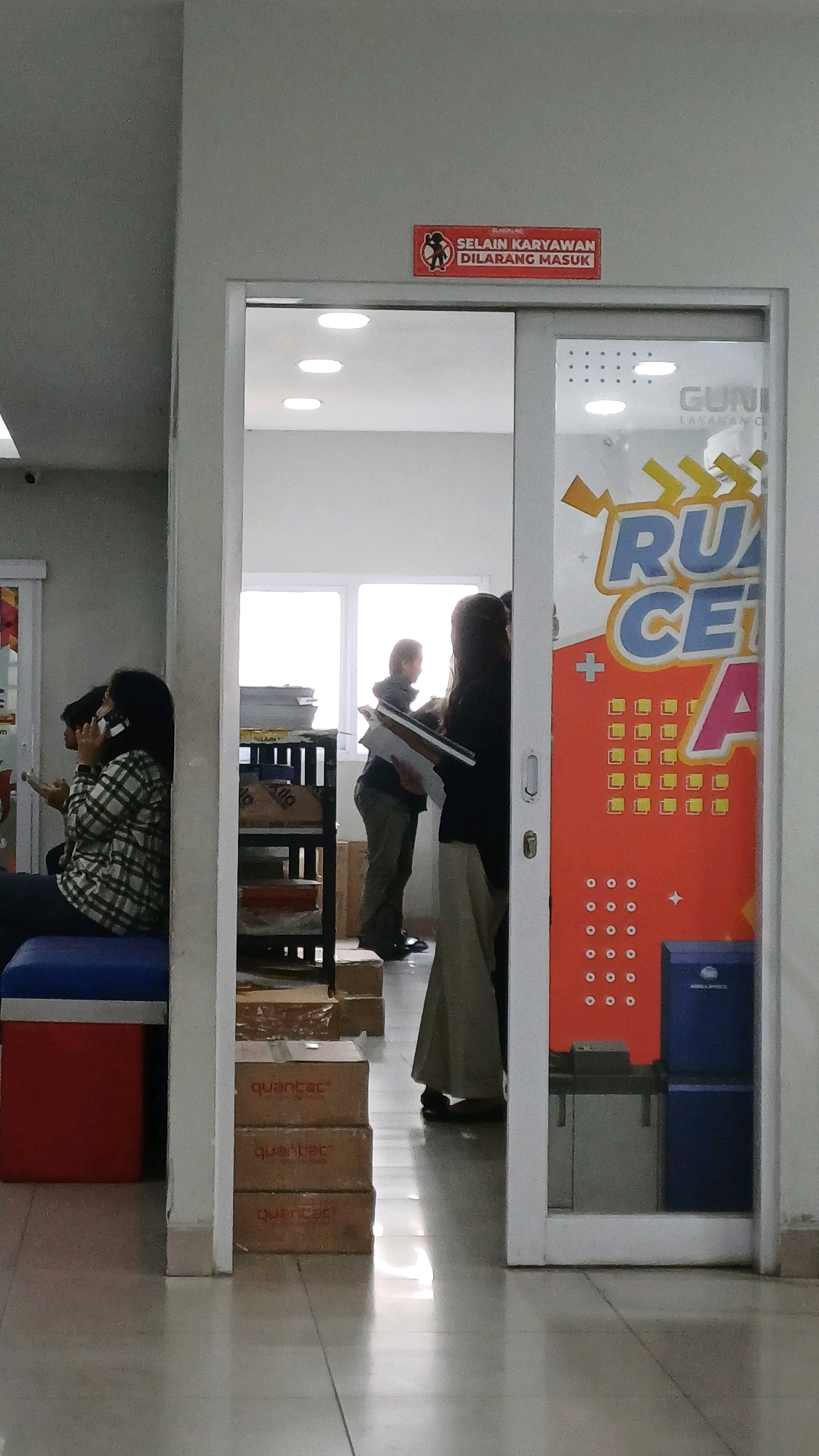 The image depicts an indoor scene, likely a reception or waiting area. Above a doorway with a sliding glass door, a red sign with white text reads "SELAIN KARYAWAN DILARANG MASUK" which translates to "OTHERS NOT ALLOWED TO ENTER".
Inside the area visible through the doorway, there are several people. On the left, a woman with dark hair is seated, talking on a mobile phone, and holding a tablet. She is wearing a checkered shirt and is positioned near a blue and red piece of furniture. To her right, inside the doorway, two individuals are standing. One, wearing a grey jacket and green pants, is looking towards a window and holding some documents. Next to them, a person wearing a dark top and a long grey skirt is also holding documents.
The environment includes a shelving unit filled with boxes, and stacks of brown cardboard boxes labeled "quantat" are visible on the floor in the foreground. The floor is tiled and reflects light. On the right side of the doorway, a large orange graphic with text and shapes is adhered to the glass. The overall lighting suggests an indoor setting with artificial lights and natural light coming from the window. The location is identified as Cibinong, Indonesia.