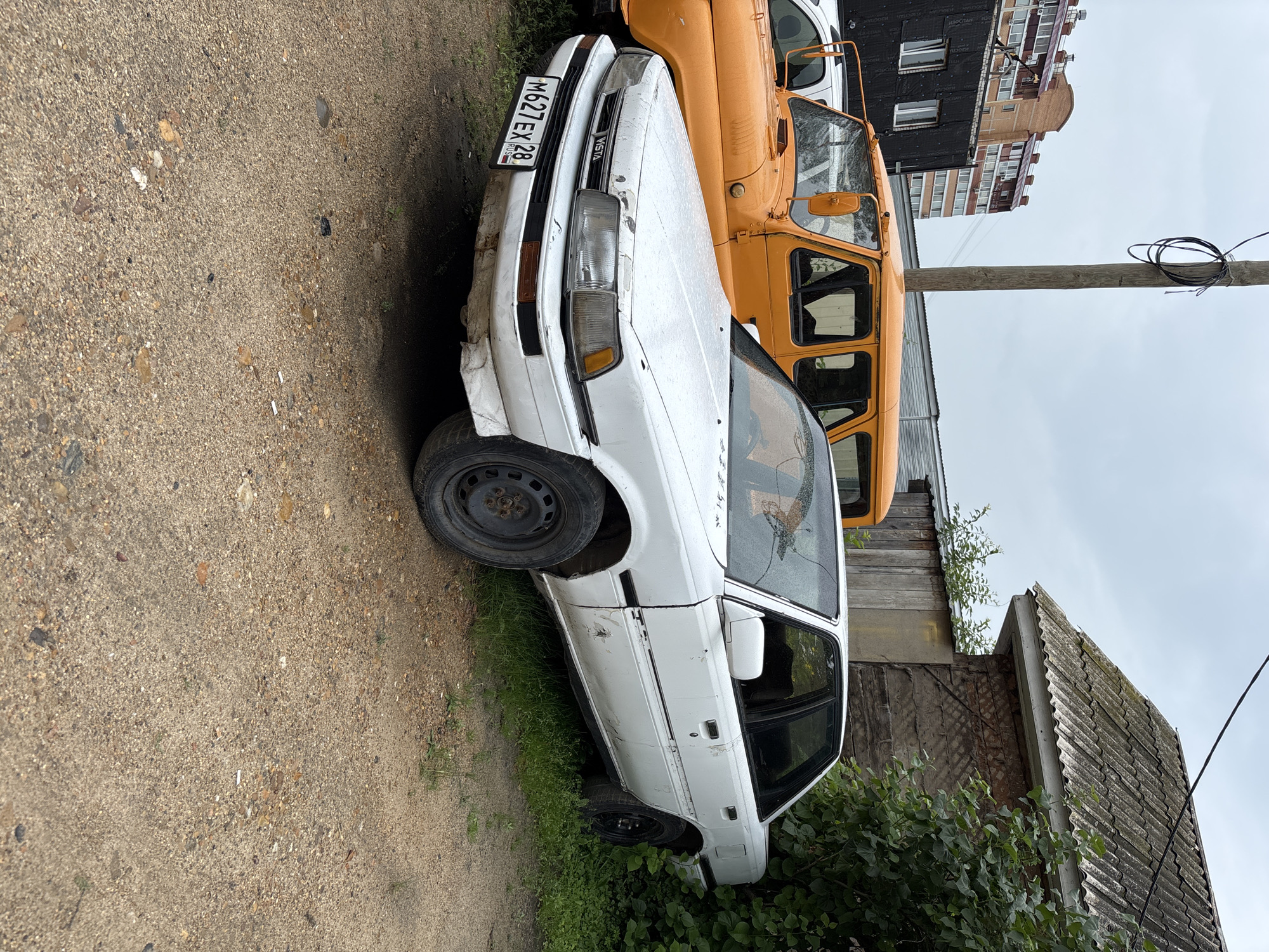 A white sedan, likely a Toyota Vista, is parked on a gravel and dirt lot in Blagoveshchensk, Russia. The car is in poor condition, with visible rust, dents, and faded paint, particularly on the front bumper and along the side panels. A black license plate with Russian characters and numbers (M627EX 28) is affixed to the front. To the left of the sedan, the front of an orange utility vehicle, possibly a UAZ, is visible. In the background, a brick apartment building with multiple stories and air conditioning units stands under a cloudy sky. To the right of the sedan, a portion of a weathered wooden building with a corrugated roof and overgrown green bushes is present. A wooden utility pole with coiled wires stands prominently in the center-left of the image. The ground is scattered with small stones, dirt, and some debris, including cigarette butts. The overall scene suggests an outdoor storage area or a less maintained part of the city.