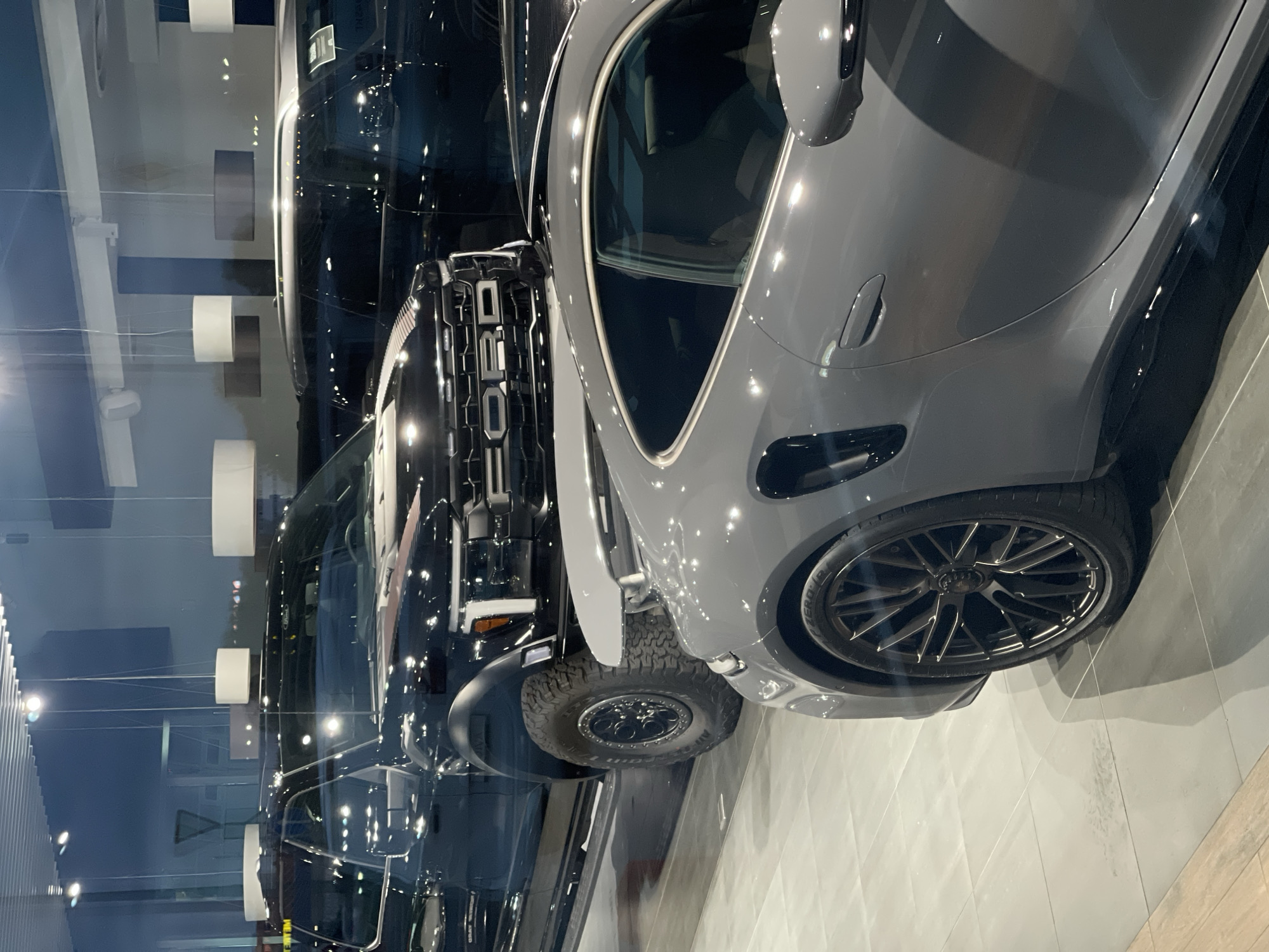 A gray Porsche 911 Turbo S is parked in the foreground, reflecting the lights of a showroom. Behind it, a black Ford F-150 Raptor pickup truck is visible, also in a showroom setting with large windows. The showroom floor is tiled, and overhead lighting fixtures are suspended from the ceiling. The image appears to have been taken at an angle from a slightly elevated position.