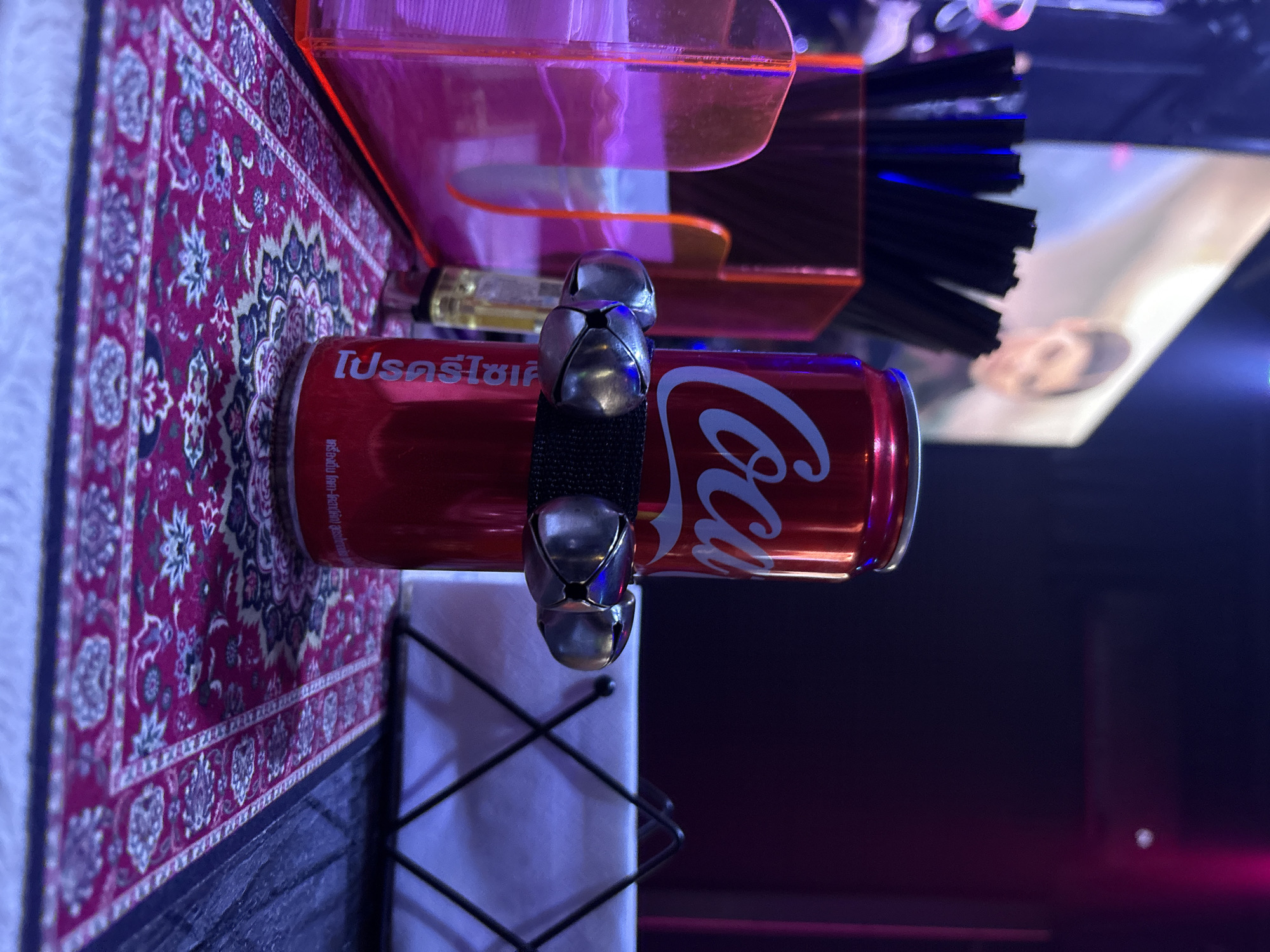 This photo depicts a Coca-Cola can adorned with jingle bells resting on a surface covered with a patterned rug. The can has text in Thai. A plastic container holding black straws is visible behind the can. In the background, there is a blurry image of a person and lighting suggests a nightclub or bar setting. Based on the Thai text, this image is likely from Baan Bang Rak, Thailand.