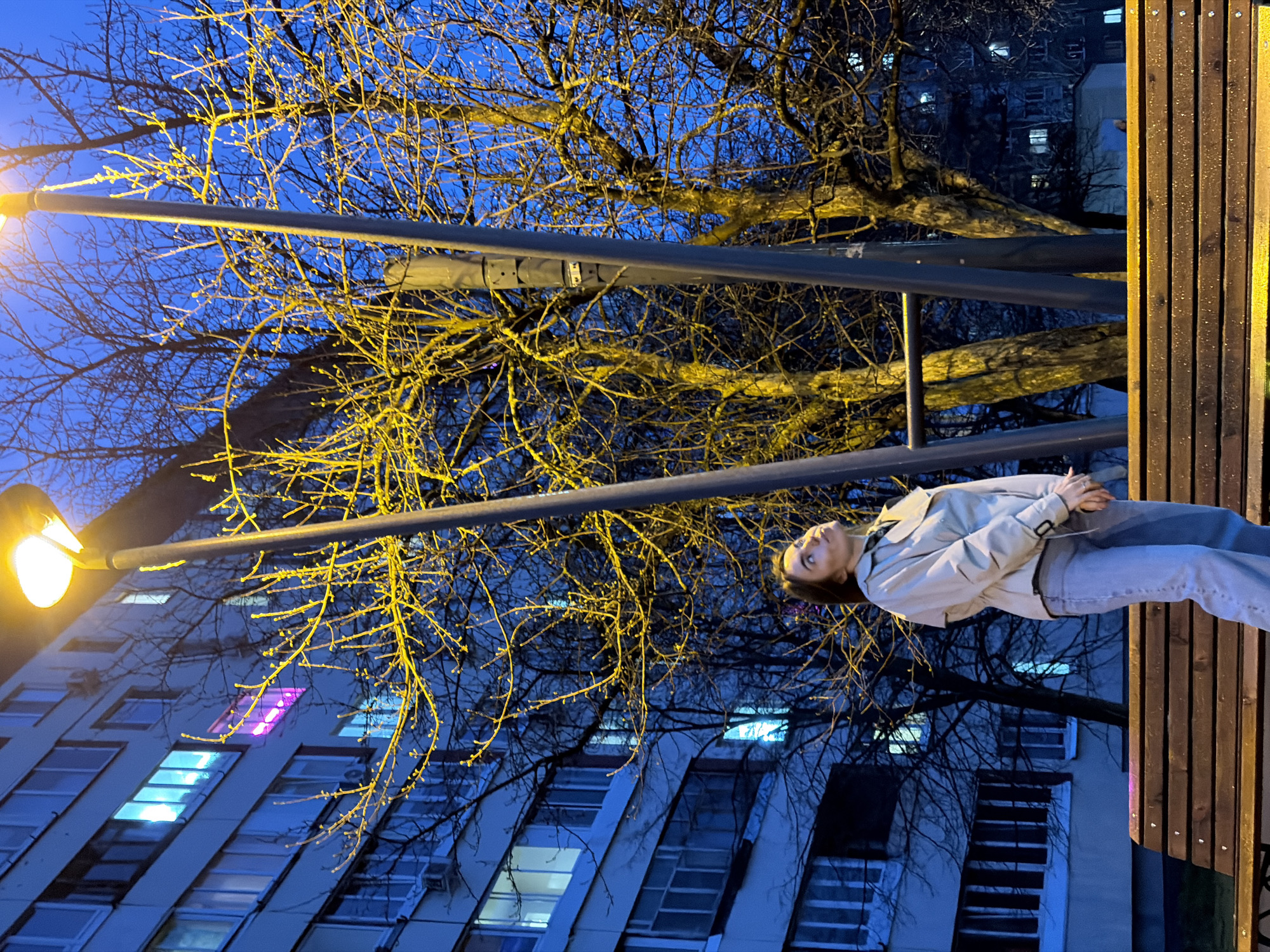 A young woman lies on a wooden bench under a street lamp in Izhevsk, Russia. Bare tree branches are silhouetted against the night sky, partially obscuring a building with multiple illuminated windows, including one window with pink light. The scene is lit by the warm glow of the street lamp, contrasting with the cool blue tones of the building and sky. The woman is wearing a light-colored jacket and jeans, her hands clasped. Metal railings run parallel to the bench.