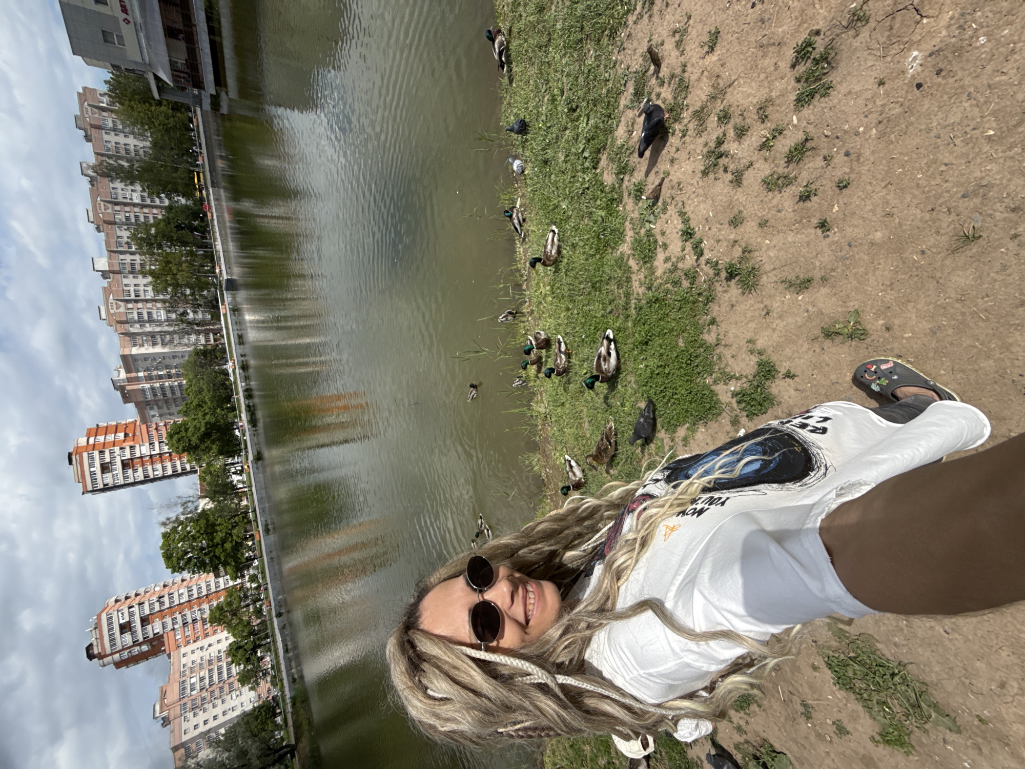 The image, taken from a first-person perspective, captures an outdoor scene by a body of water. A smiling person with long blonde dreadlocks or braids and round sunglasses occupies the right side of the frame, looking up at the camera. They are wearing a white t-shirt with a dark graphic design that includes partially visible text, "NOW YOUR," and dark sandals adorned with colorful charms.
To the person's left, a grassy bank slopes down to a calm body of water, likely a pond or canal. Numerous ducks, primarily mallards, are visible both in the water and on the grassy edge. Several pigeons are also present on the dirt path and grassy areas.
In the background, on the far side of the water, a line of tall, multi-story residential or commercial buildings with varied facades stands against a partly cloudy sky. Trees are interspersed along the waterfront, between the buildings and the water. The setting appears to be an urban or suburban park or waterfront promenade during bright daylight hours. The person's expression conveys a sense of happiness or enjoyment.