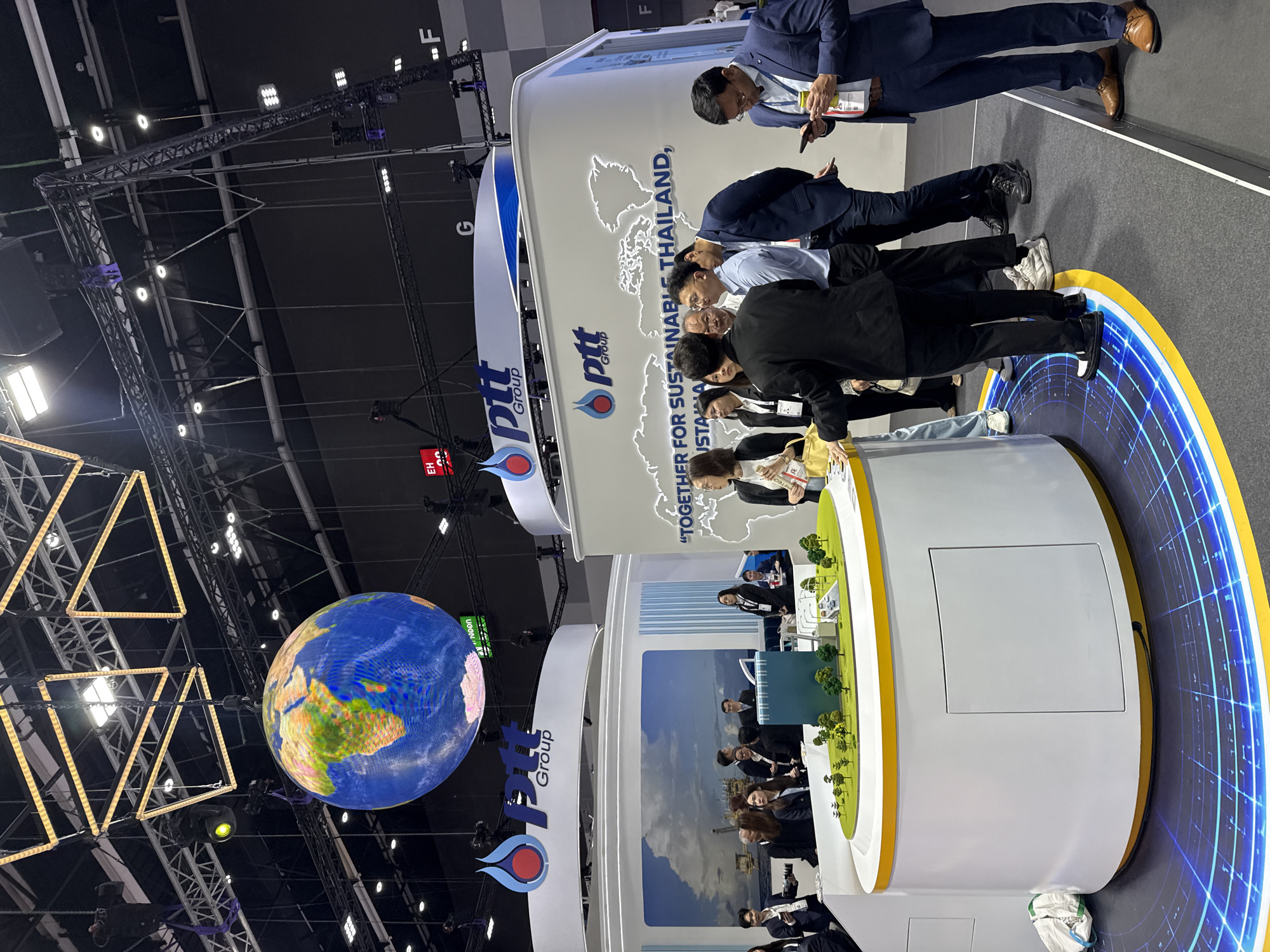 The image depicts an indoor exhibition or conference hall, likely at the IMPACT Exhibition and Convention Center in Bangkok, Thailand, given the PTT Group branding which is a major Thai energy company. In the foreground, a group of people, predominantly men in business attire, are gathered around a circular display. This display features a miniature model of a city or industrial park, complete with green landscaping, buildings, and roads with toy vehicles. The display is illuminated from below with a blue and yellow light pattern on the floor. Above, a large, illuminated globe of the Earth is suspended from the ceiling. Behind the people and the display, a large white structure with the PTT Group logo and a world map graphic is visible. Further in the background, exhibition booths and signage are dimly lit. The overall atmosphere suggests a corporate event or trade show focused on the PTT Group's operations and sustainability initiatives.