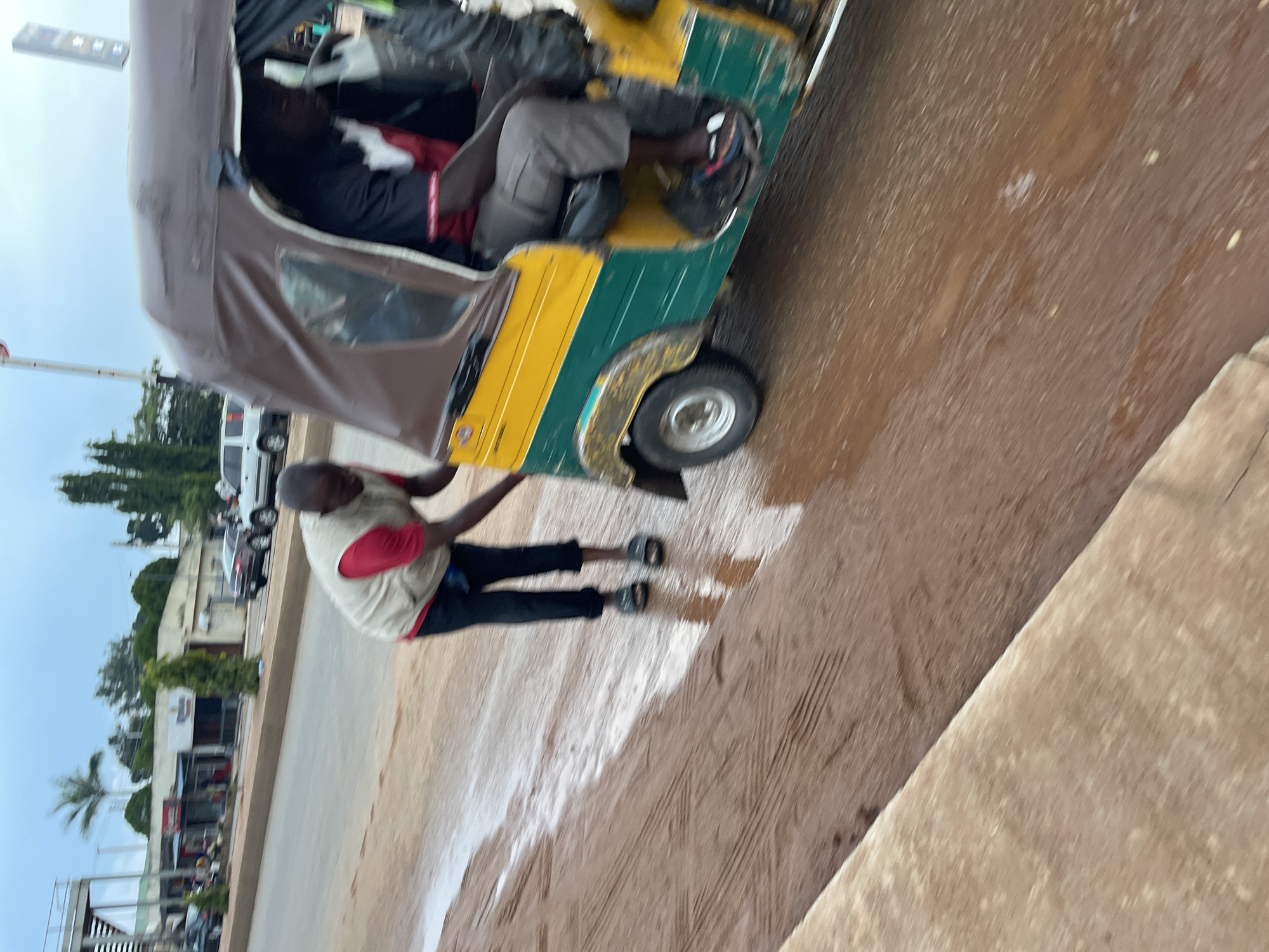 The image depicts a muddy road in Jos, Nigeria. In the foreground, a man wearing a light-colored vest over a red shirt and black trousers is pushing a yellow and green auto-rickshaw. The auto-rickshaw has a brown canopy and appears to be stuck or struggling in the mud. Another person is visible inside the auto-rickshaw, operating the vehicle. The road surface is covered in a thick layer of brown mud and water, with tire tracks visible. In the background, buildings, trees, and several vehicles, including cars and another auto-rickshaw, are present on the side of the road. The sky is overcast, suggesting cloudy weather. The scene captures a moment of difficulty or effort related to transportation on a poorly maintained road.