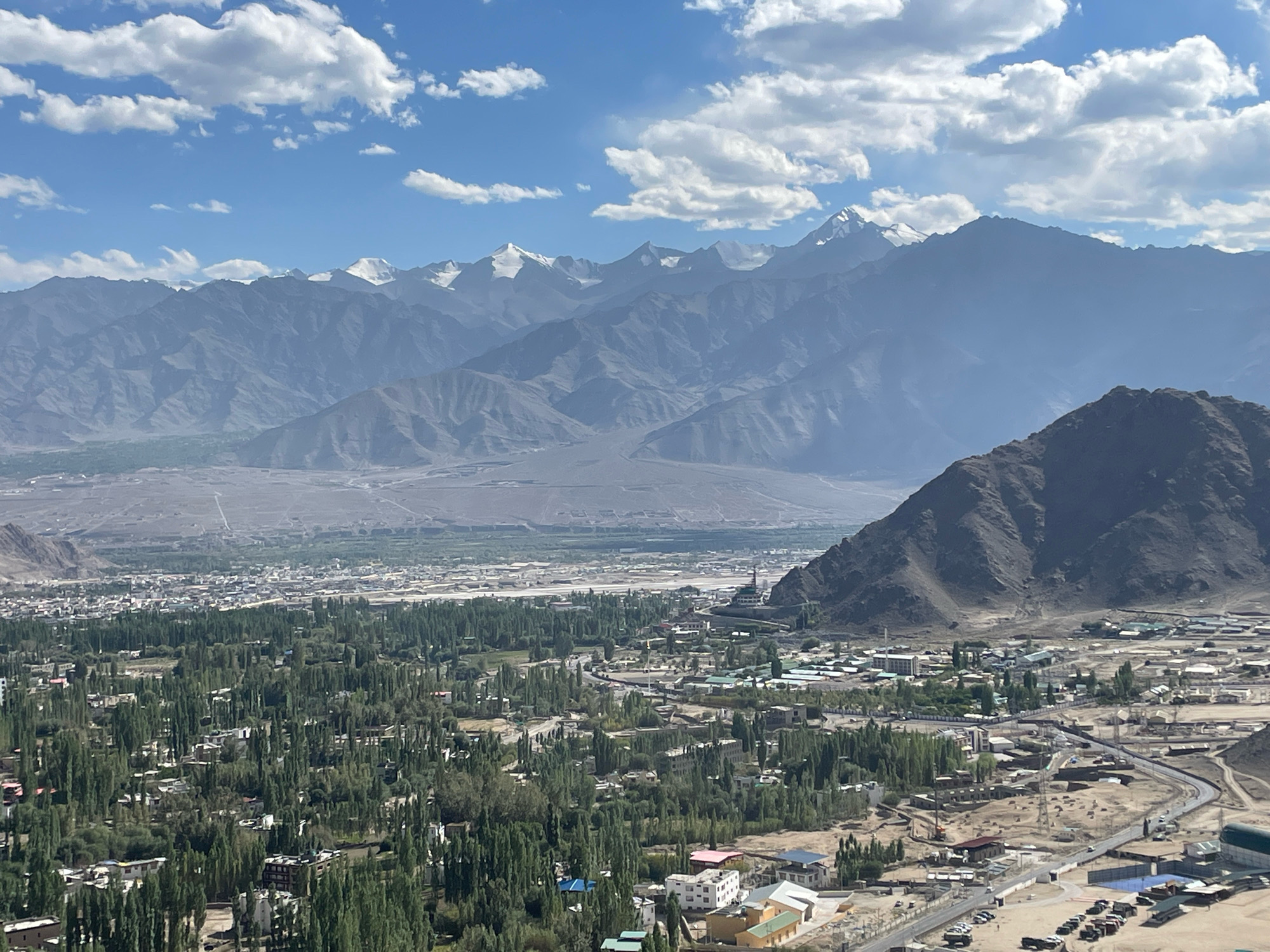 This is an aerial shot of the city of Leh, India, with the Himalayas in the background. The city is spread out in a valley, with a dense collection of buildings and trees in the foreground. The buildings are a mix of residential and commercial structures, with some larger institutional buildings visible. The trees are primarily deciduous, forming a green canopy over much of the city. In the distance, a range of rugged, snow-capped mountains rises sharply against a bright blue sky dotted with white clouds. One prominent white stupa, or Buddhist monument, is visible within the city, near the base of a steep, barren hill. The overall impression is one of a bustling urban center nestled within a dramatic natural landscape.