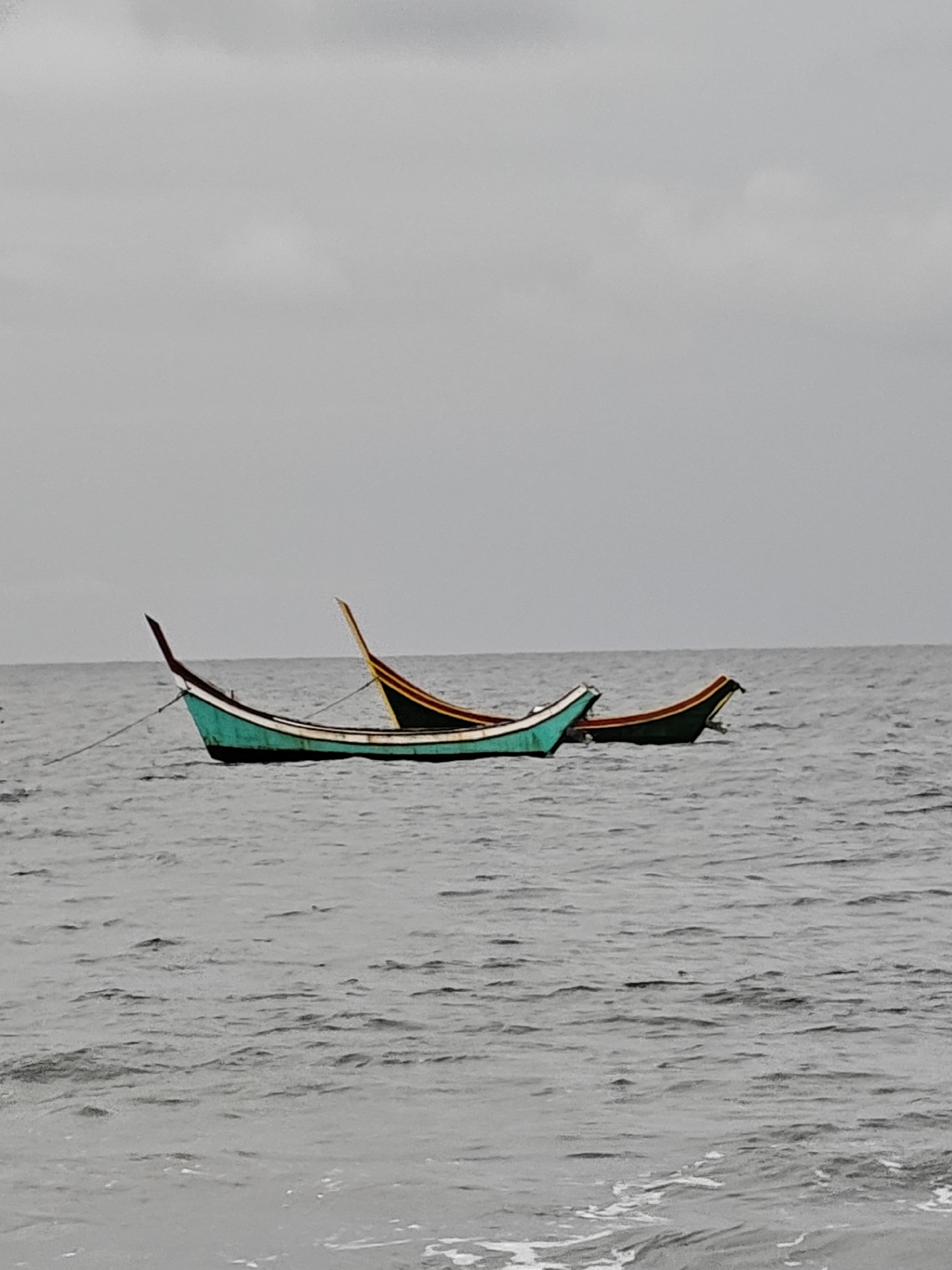 Two traditional Indonesian fishing boats, one teal and white, the other dark green and yellow, are moored on a calm, gray sea under an overcast sky. The teal boat is in the foreground, tethered by a rope to an unseen anchor point. The dark green boat is positioned slightly behind and to the right of the teal boat, also on the water. The environment is serene, with gentle ripples on the water's surface and a subdued, diffused light. The scene suggests a quiet moment in a coastal area, possibly the waters off Curehtunong, Indonesia, where such boats are common.