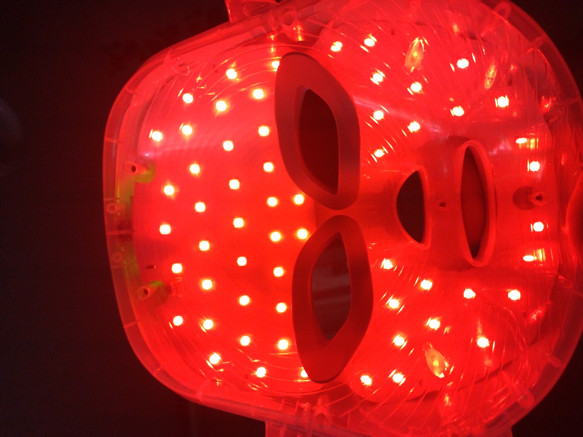 An LED facial mask glows red against a dark background. The mask is made of clear plastic with numerous small red LED lights embedded in its surface. It has cutouts for the eyes and mouth, with padded rims. The mask appears to be a single object, not worn by anyone, and is centered in the frame. The lighting is dim and casts a red hue over the entire object. The location is unknown.
